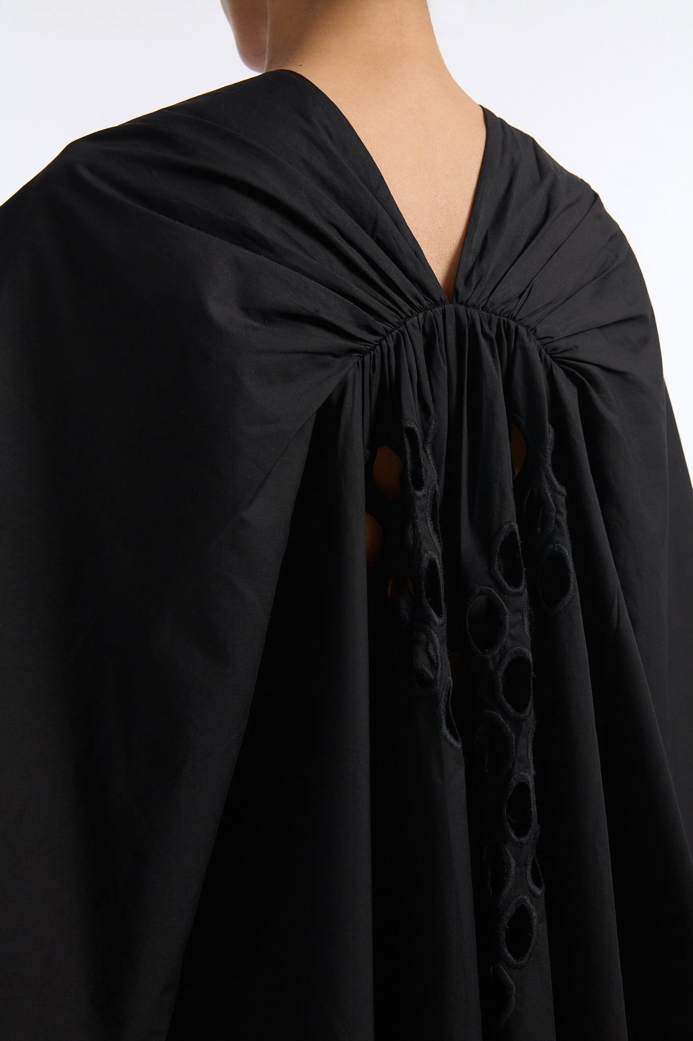 Female model with slicked back black hair in a bun wears black cotton V-neck caftan with rounded diamond cutouts along chest and upper abdomen as well as on the back. The caftan covers both arms and has a shorter front than back with slits along either side. 