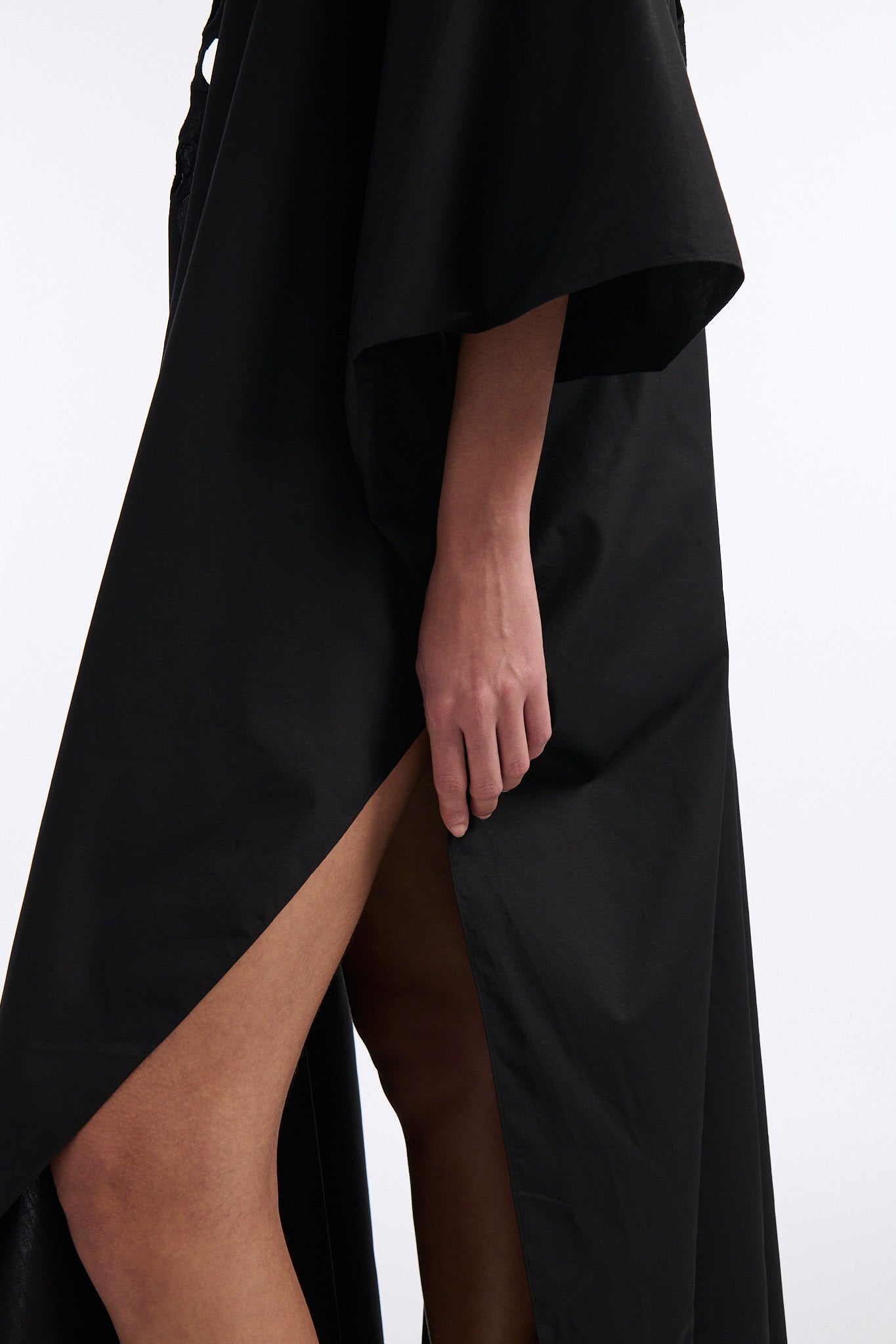 Female model with slicked back black hair in a bun wears black cotton V-neck caftan with rounded diamond cutouts along chest and upper abdomen as well as on the back. The caftan covers both arms and has a shorter front than back with slits along either side. 