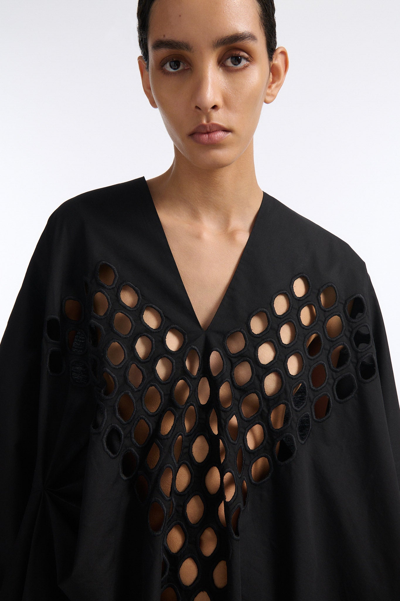 Female model with slicked back black hair in a bun wears black cotton V-neck caftan with rounded diamond cutouts along chest and upper abdomen as well as on the back. The caftan covers both arms and has a shorter front than back with slits along either side. 