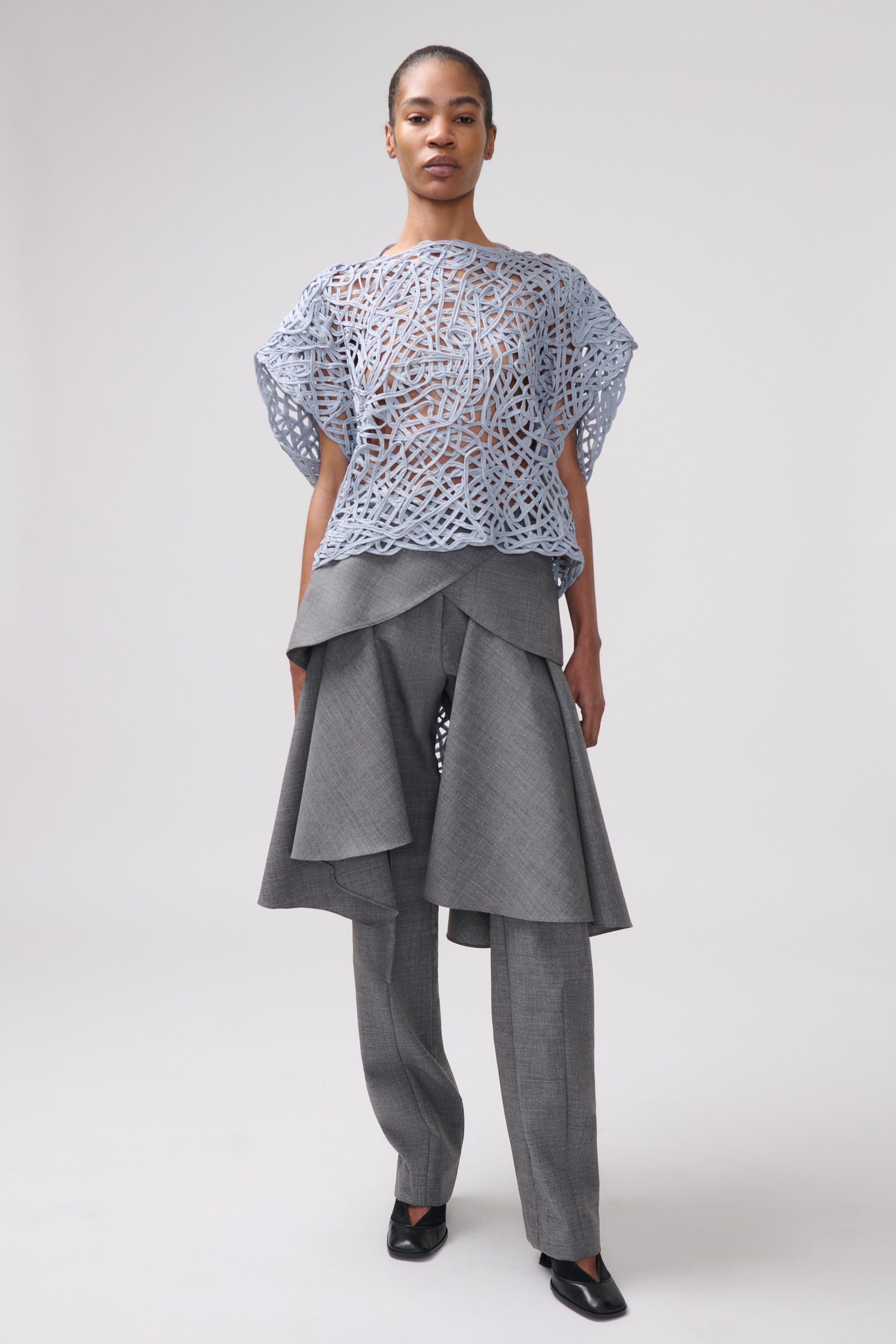 A model stands against a light gray studio background wearing a light blue sheer top  with an abstract pattern, layered over a gray asymmetrical draped gray trousers.