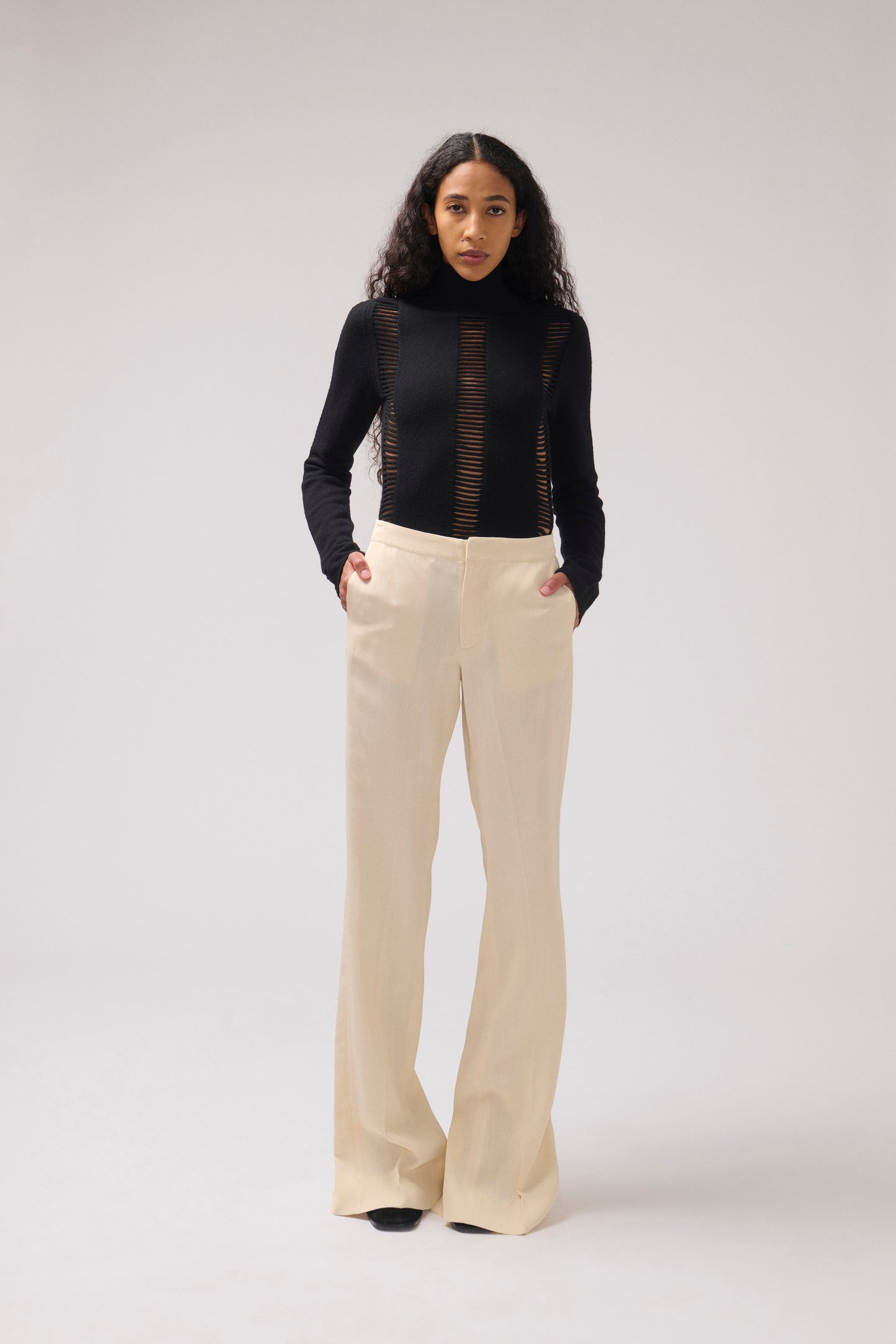 A model stands against a light gray studio background wearing a black long-sleeve mock-neck top with sheer vertical mesh panels and high-waisted, wide-leg cream pants.