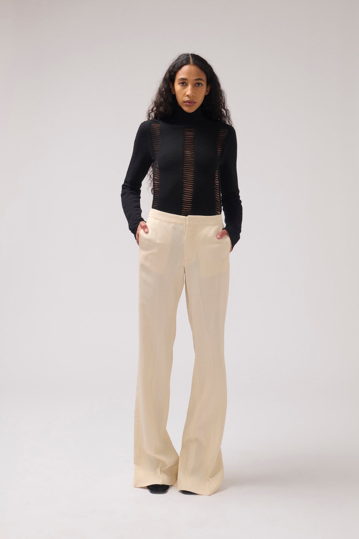 A model stands against a light gray studio background wearing a black long-sleeve mock-neck top with sheer vertical mesh panels and high-waisted, wide-leg cream pants.