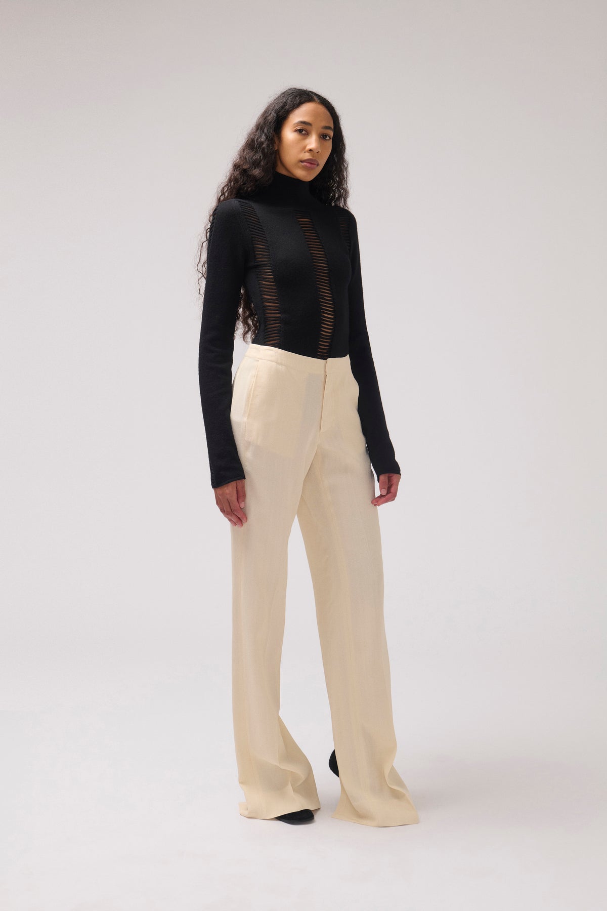 A model stands against a light gray studio background wearing a black long-sleeve mock-neck top with vertical sheer cutout details on the front and high-waisted, wide-leg cream trousers.