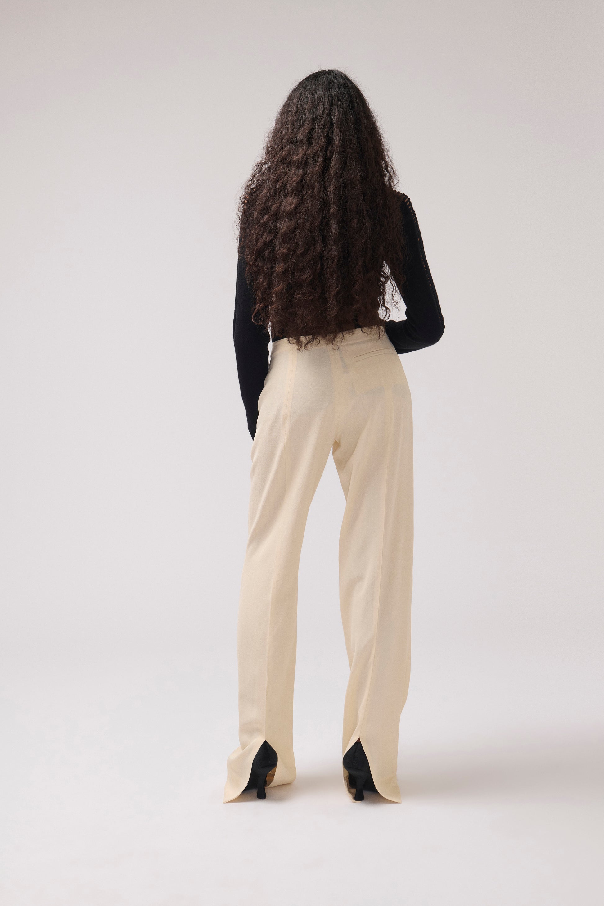 A woman with long, dark curly hair is shown from behind wearing cream-colored wide-leg trousers, a black long-sleeve top, and black heels against a plain light gray studio background.