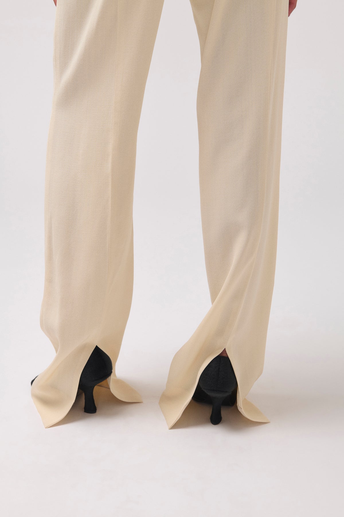 Close-up back view of a person wearing cream-colored wide-leg trousers with split hems and black kitten heels, shown against a plain light gray studio background.