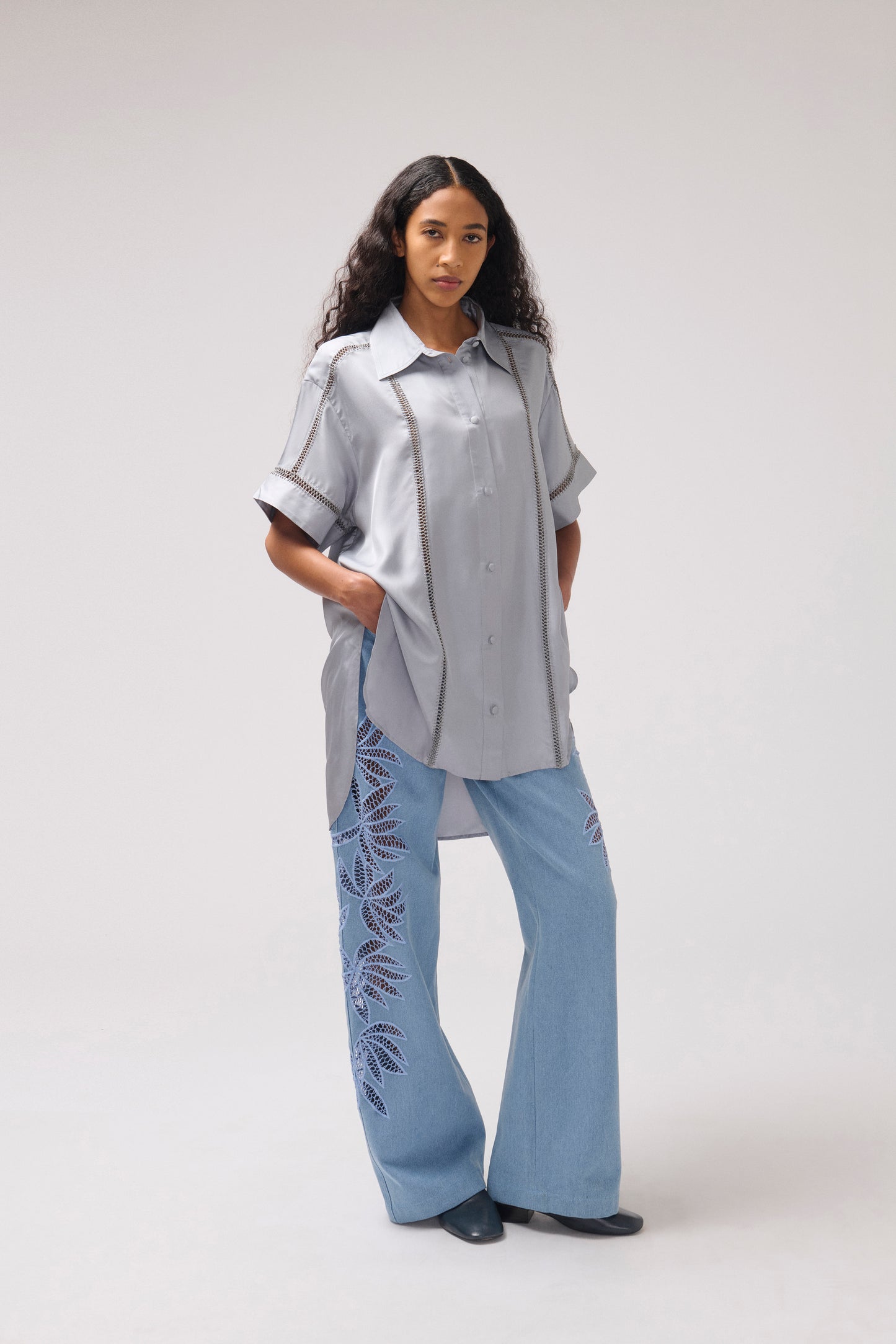 A model stands against a light gray studio background wearing a light gray short-sleeve button-up shirt with dark contrast piping and light blue wide-leg pants featuring dark blue leaf embroidery.