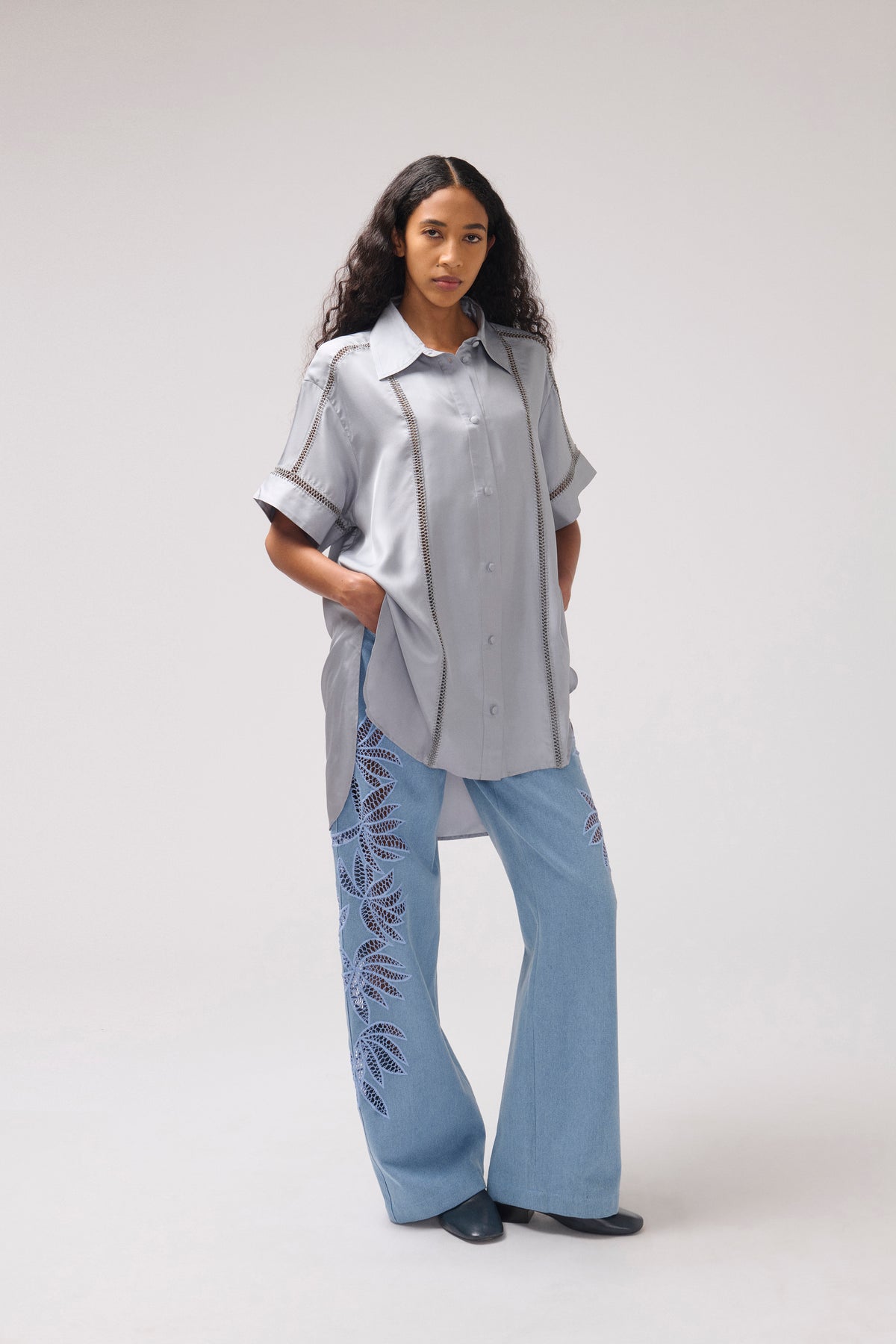 A model stands against a light gray studio background wearing a light gray short-sleeve button-up shirt with dark contrast piping and light blue wide-leg pants featuring dark blue leaf embroidery.