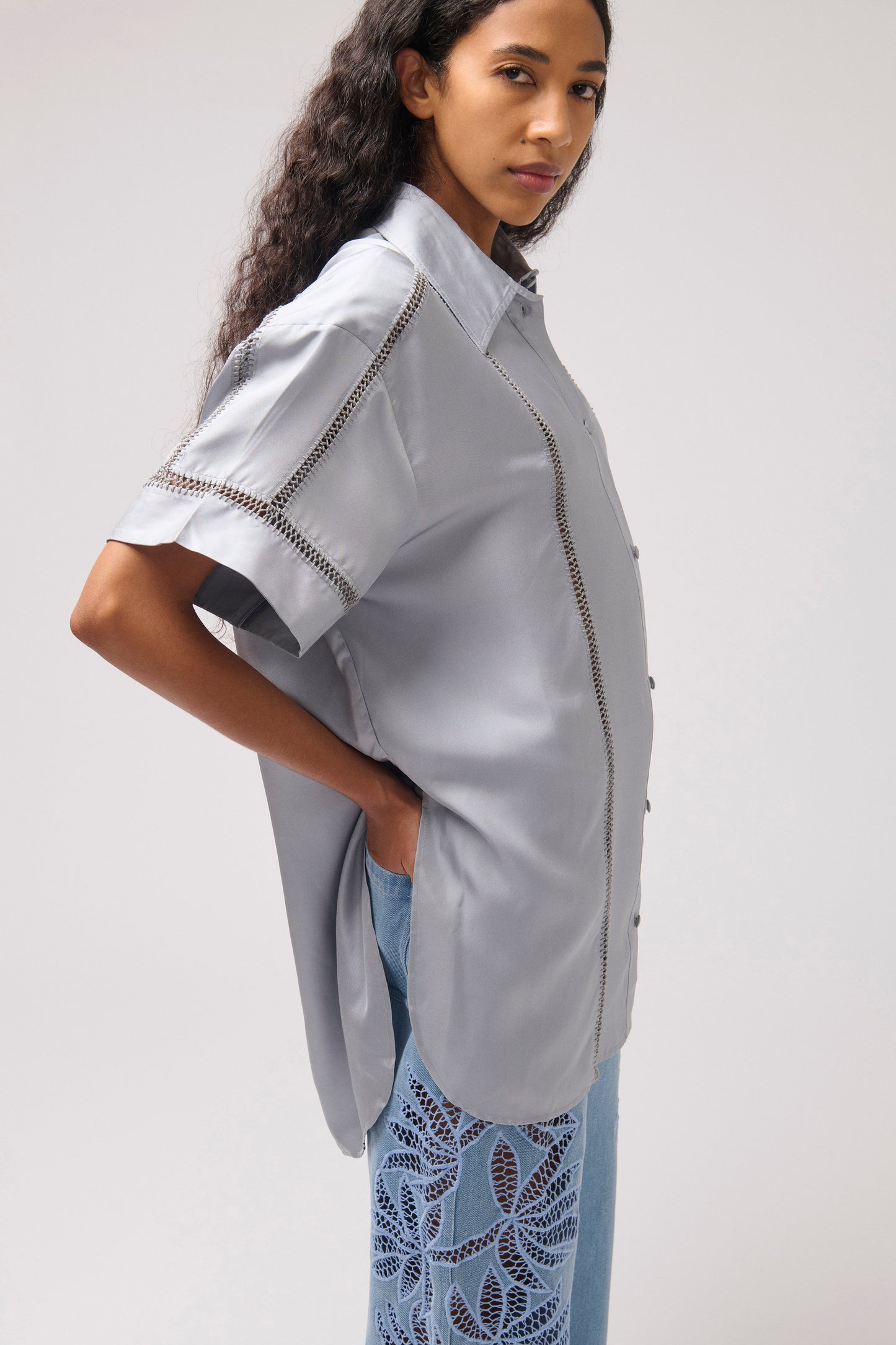 A woman wearing a light gray short-sleeve button-up shirt with decorative chain trim details, paired with light blue patterned pants, photographed against a neutral light gray studio background.