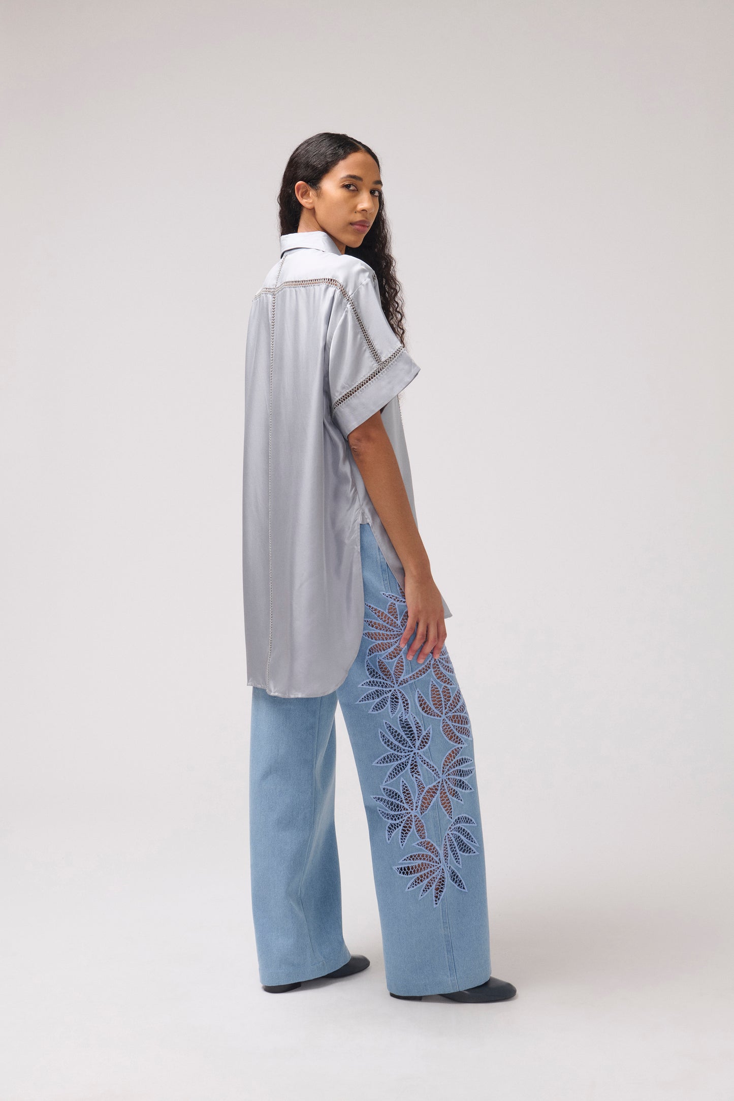 A model is shown from a side-back angle wearing a light gray short-sleeve button-up shirt and light blue wide-leg jeans featuring purple and blue floral embroidery down one leg, set against a plain light gray studio background.