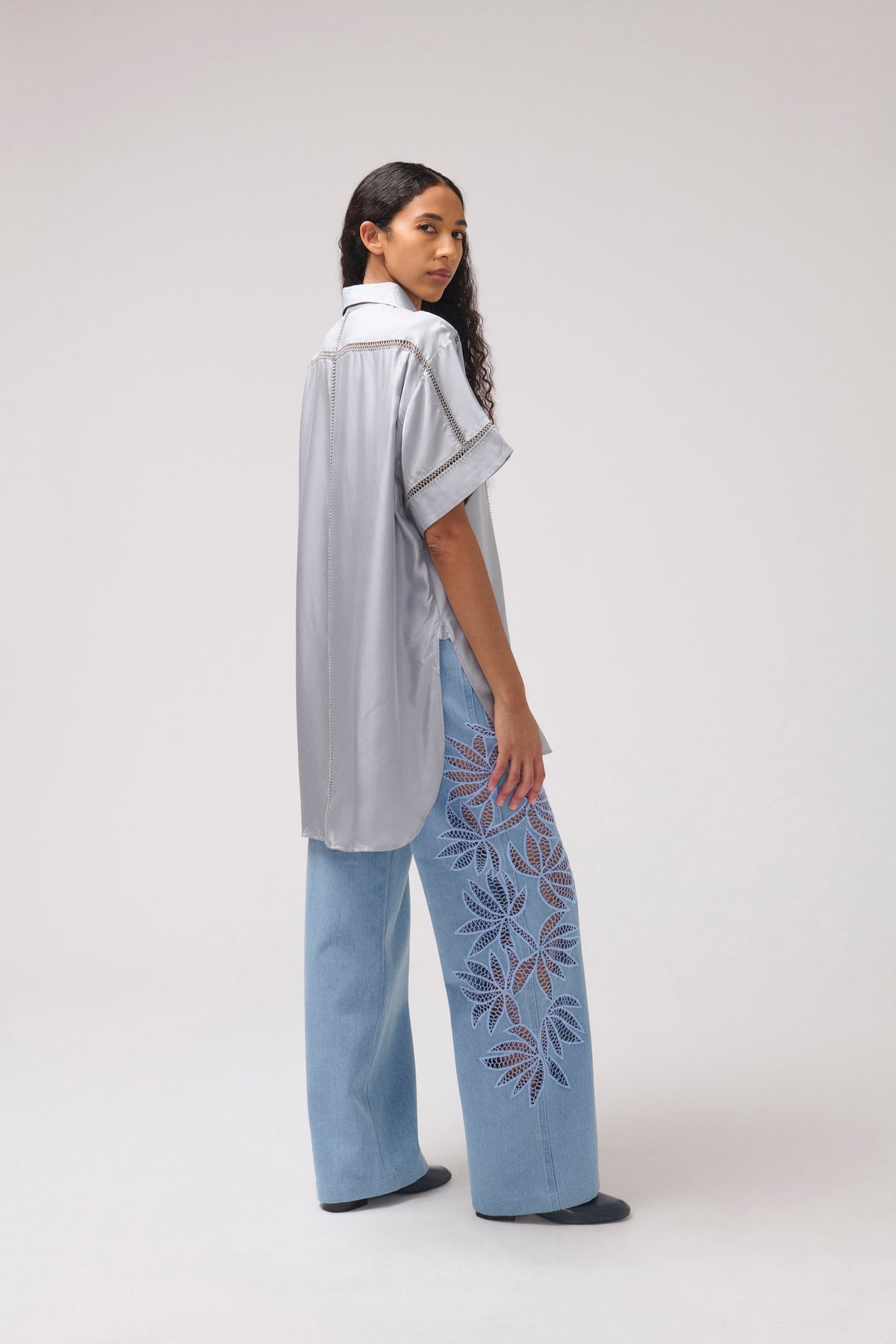 A model is shown from a side-back angle wearing a light gray short-sleeve button-up shirt and light blue wide-leg jeans featuring purple and blue floral embroidery down one leg, set against a plain light gray studio background.