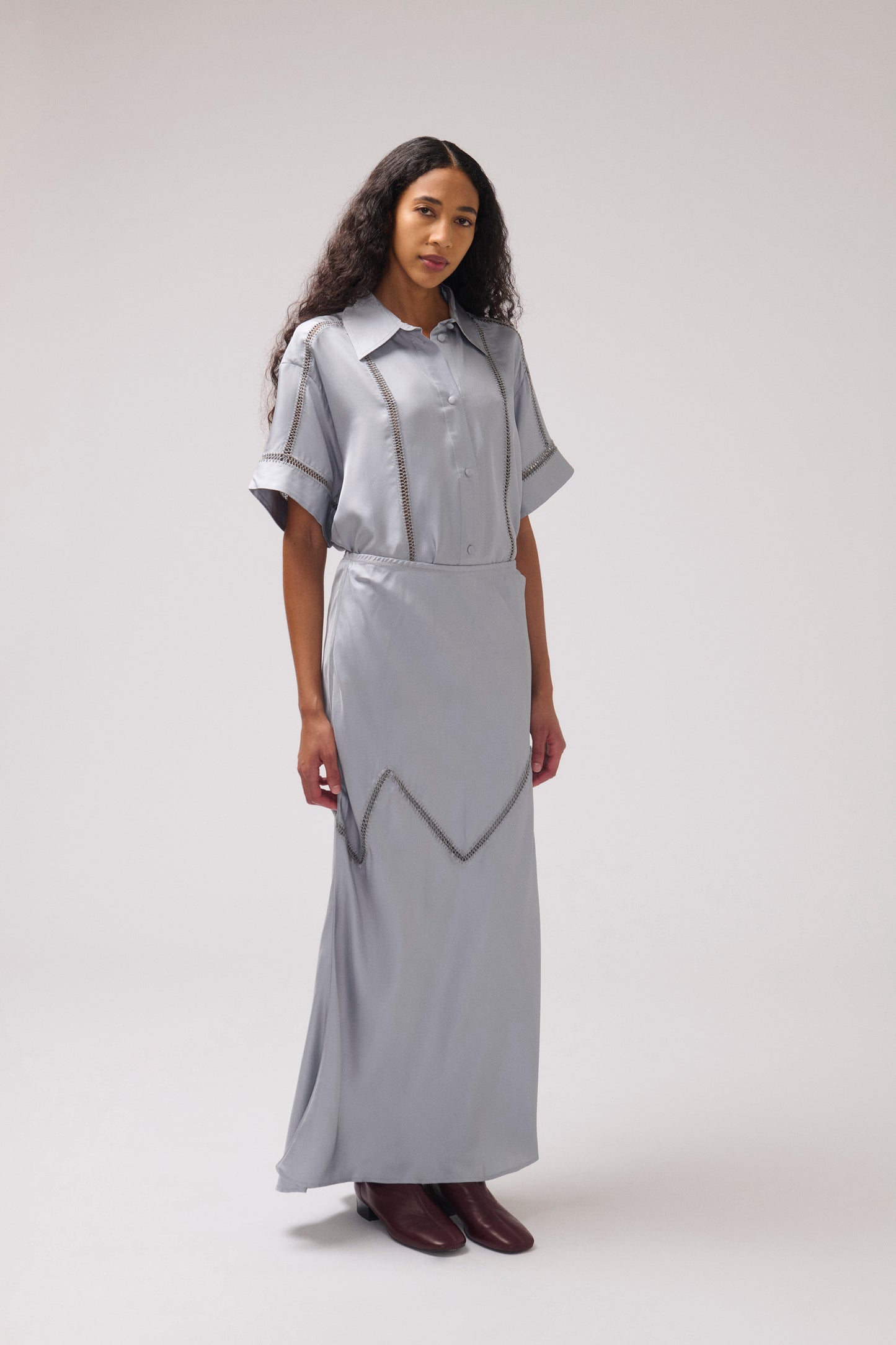 A model wearing a light gray short-sleeve shirt dress with silver metallic geometric trim, standing against a neutral light gray studio background.