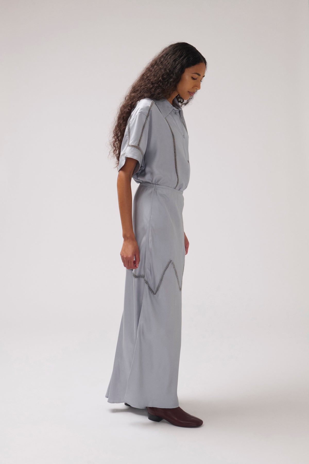 A model wearing a light gray midi dress with short flutter sleeves, a high collar, and a zigzag pattern on the skirt, standing against a plain light gray studio background.