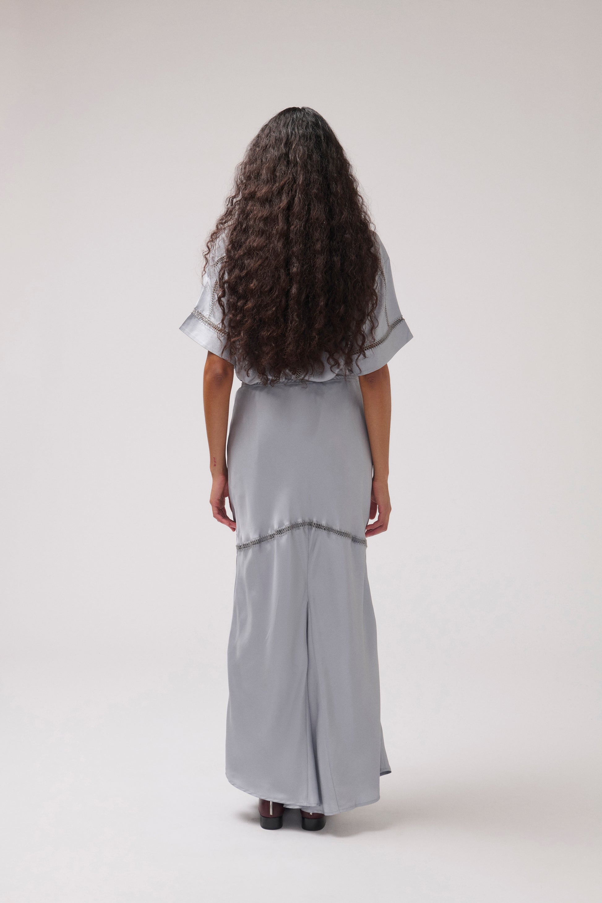 A woman with long, curly dark brown hair is shown from behind wearing a light gray, short-sleeved maxi dress with a tiered skirt. She is standing against a plain, light gray studio background.