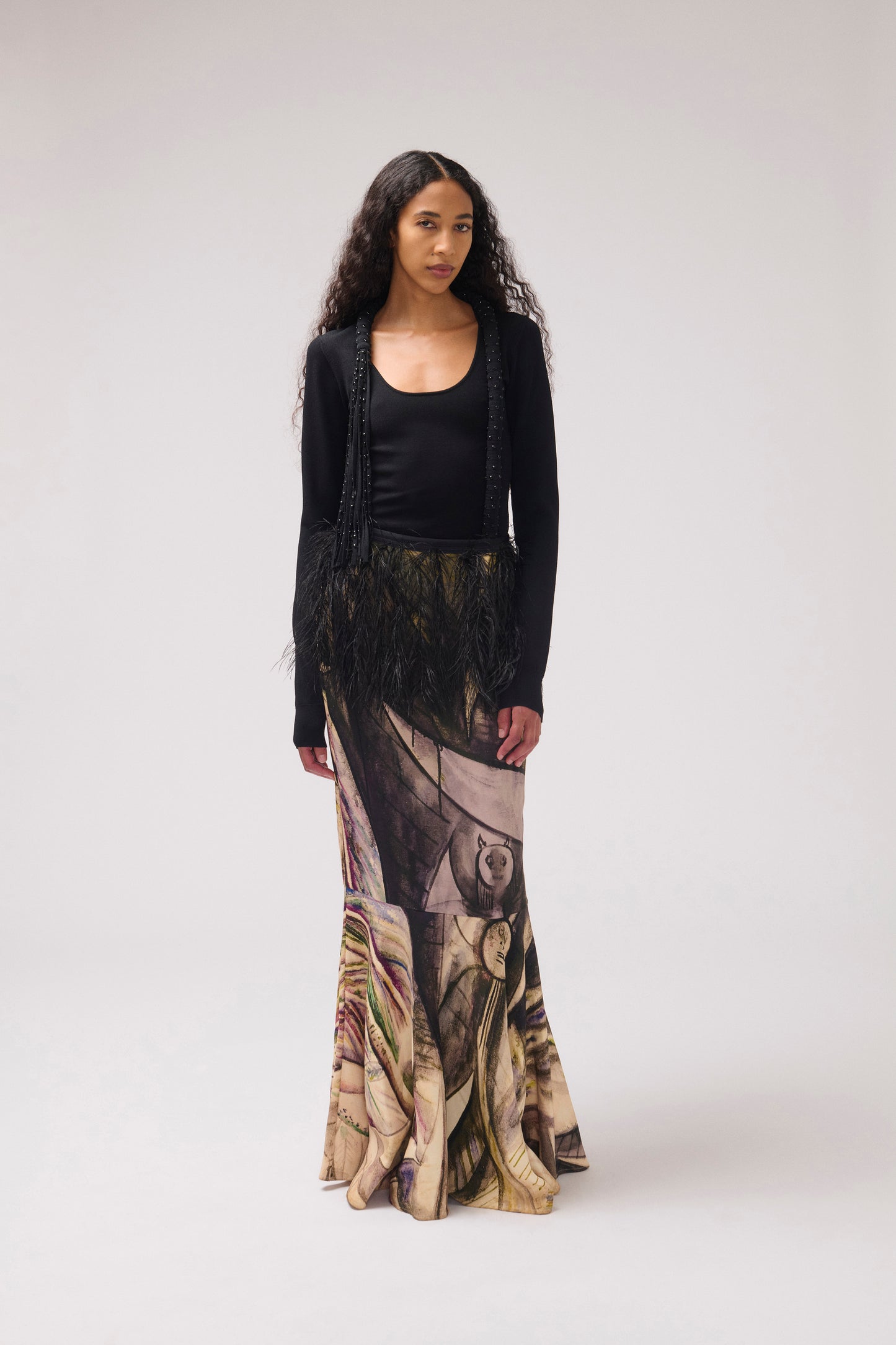 A model stands against a light gray studio background wearing a black top and a long, wide skirt featuring a painterly print with feather detail at the waist.