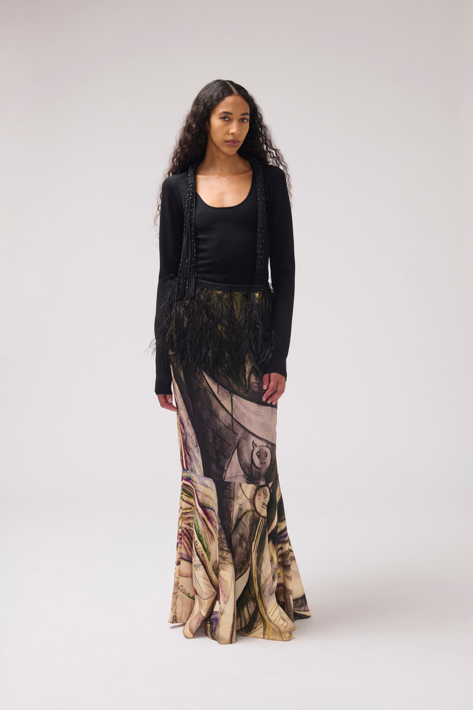 A model stands against a light gray studio background wearing a black top and a long, wide skirt featuring a painterly print with feather detail at the waist.