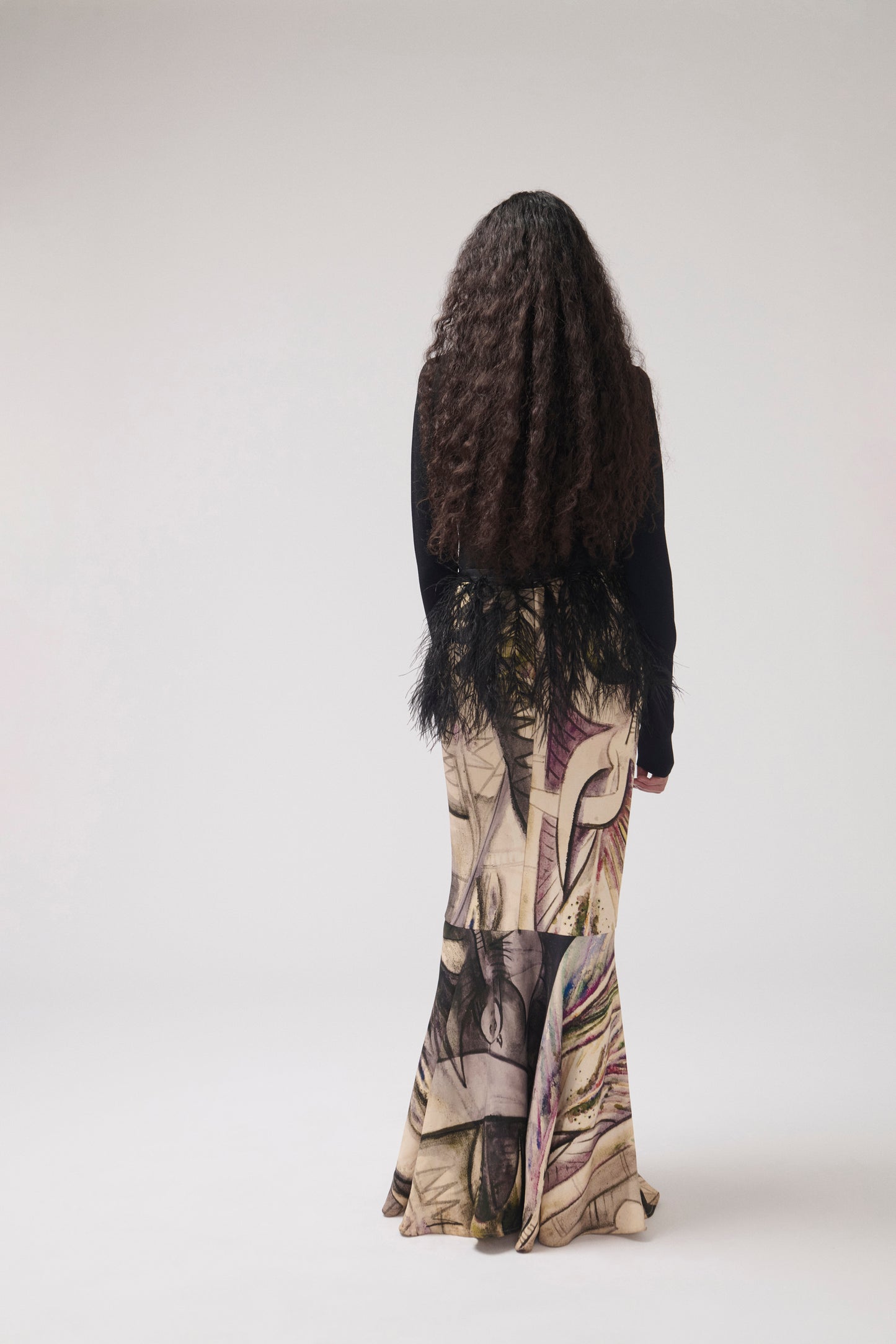 The back of a model who stands against a light gray studio background wearing a black top and a long, wide skirt featuring a painterly print with feather detail at the waist.