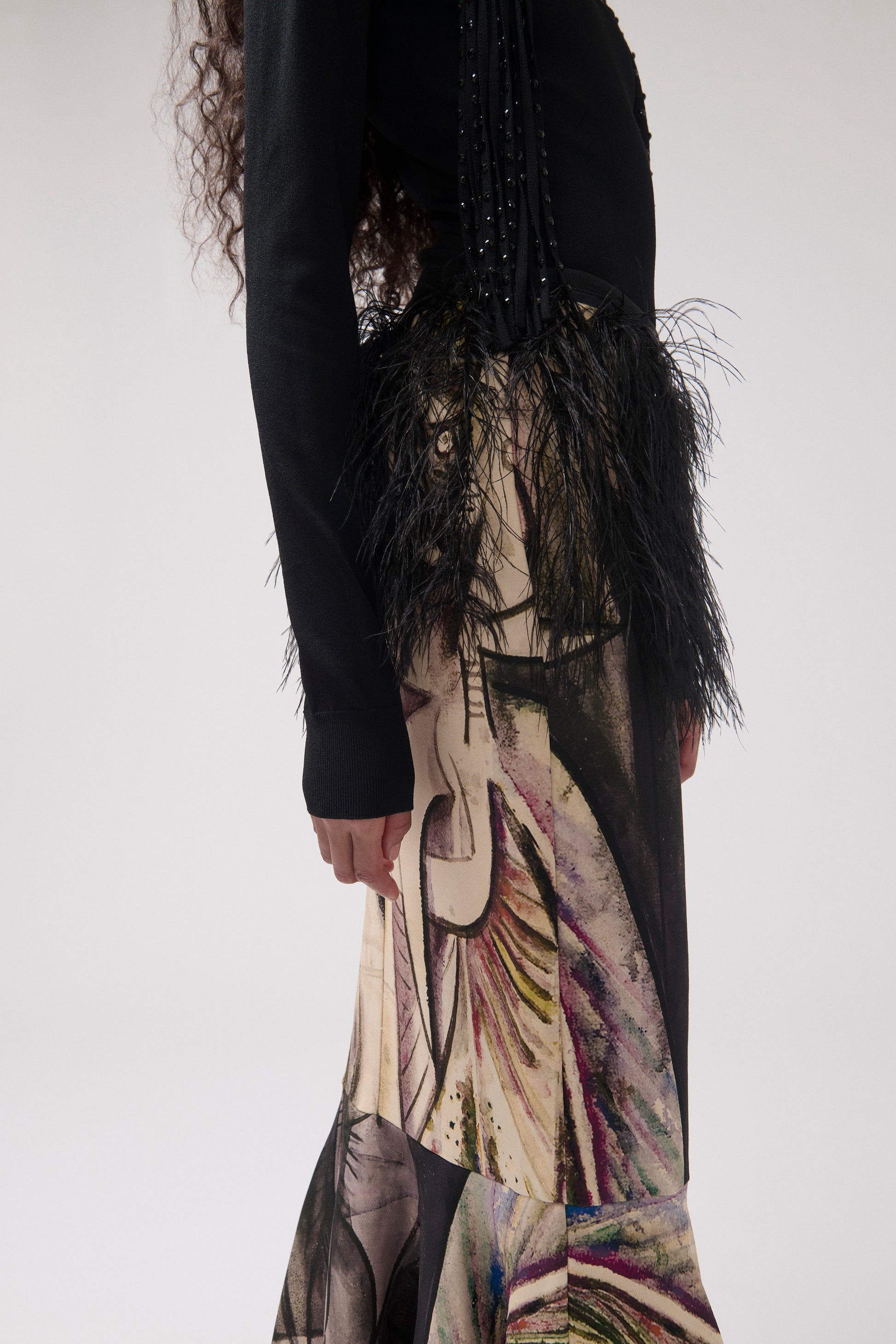 Close-up of a model standing against a light gray studio background wearing a black top with fringe detail and a long, wide skirt featuring a painterly print with feather detail at the waist.