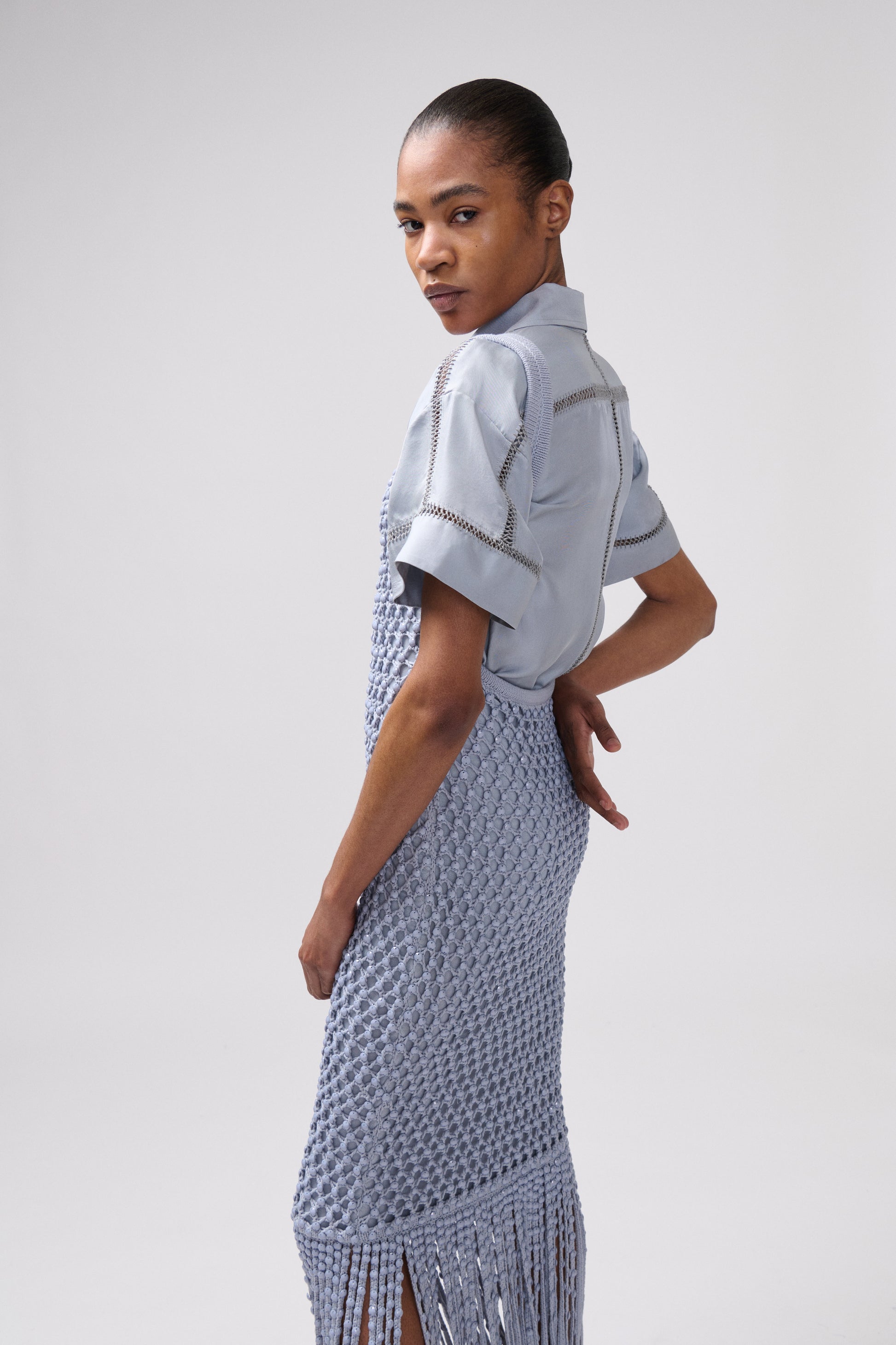 A model wearing a light blue short-sleeve top layered underneath a textured dress, photographed against a neutral light gray studio background.