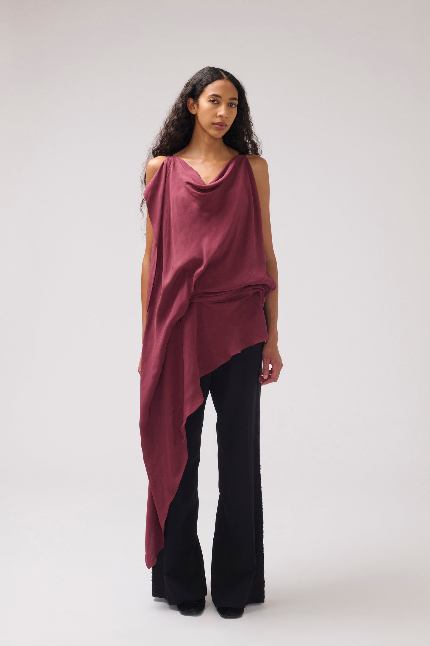 A model stands against a light gray studio background wearing a draped, asymmetrical cowl-neck top in a deep burgundy or maroon shade paired with black wide-leg pants.