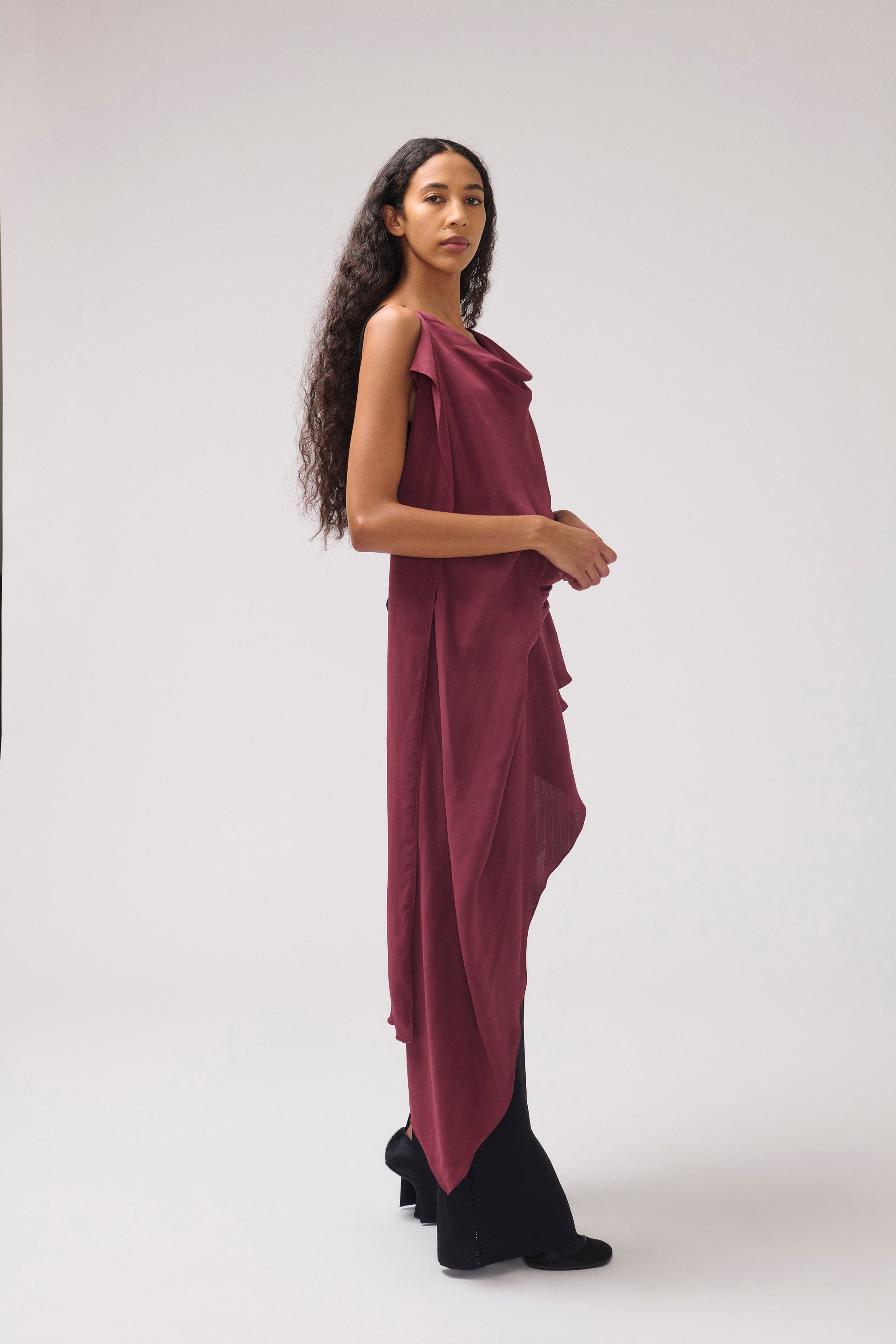 A woman modeling a draped, one-shoulder maxi dress in deep burgundy, paired with black wide-leg trousers, standing against a neutral light gray studio background.