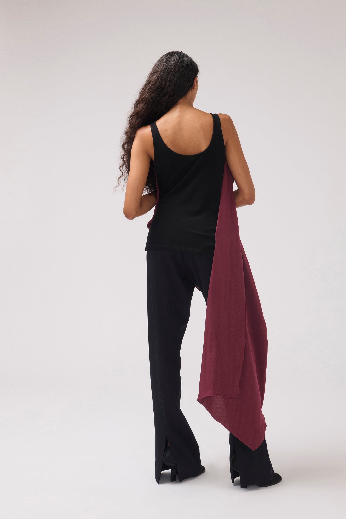 A woman is shown from behind wearing a black tank top and black wide-leg pants, holding a draped burgundy scarf or shawl against a plain light gray studio background.