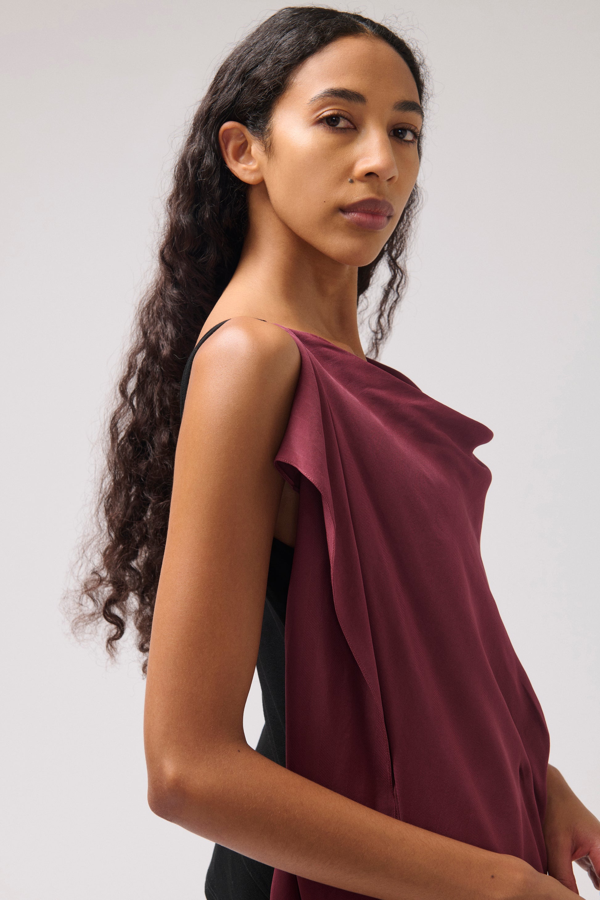 A woman modeling a draped, one-shoulder top in deep burgundy, shown from a side profile against a light gray studio background.