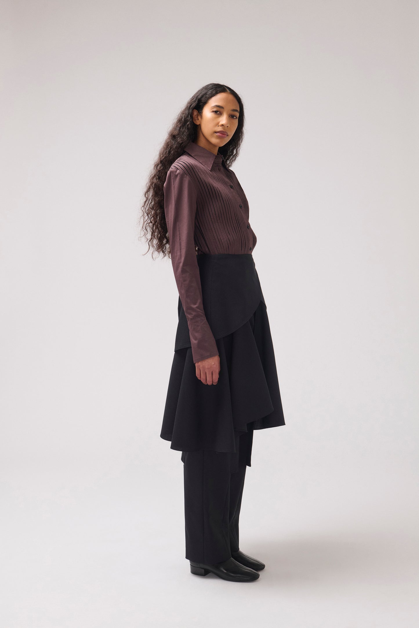 A model stands in profile wearing a dark brown ribbed long-sleeve mock-neck top, a black asymmetrical layered skirt, and black wide-leg trousers against a light gray studio background.