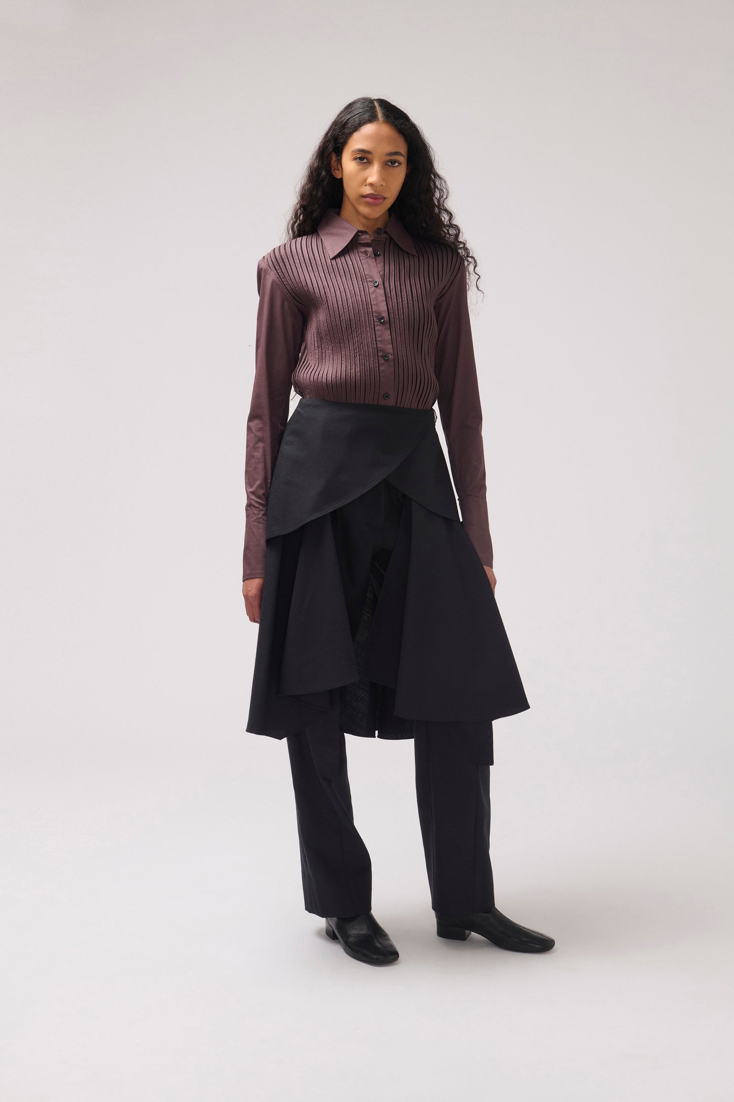 A model stands against a light gray studio background wearing a long-sleeve button-front shirt in a dark mauve-brown ribbed fabric, a black asymmetrical wrap skirt, black trousers, and black shoes.