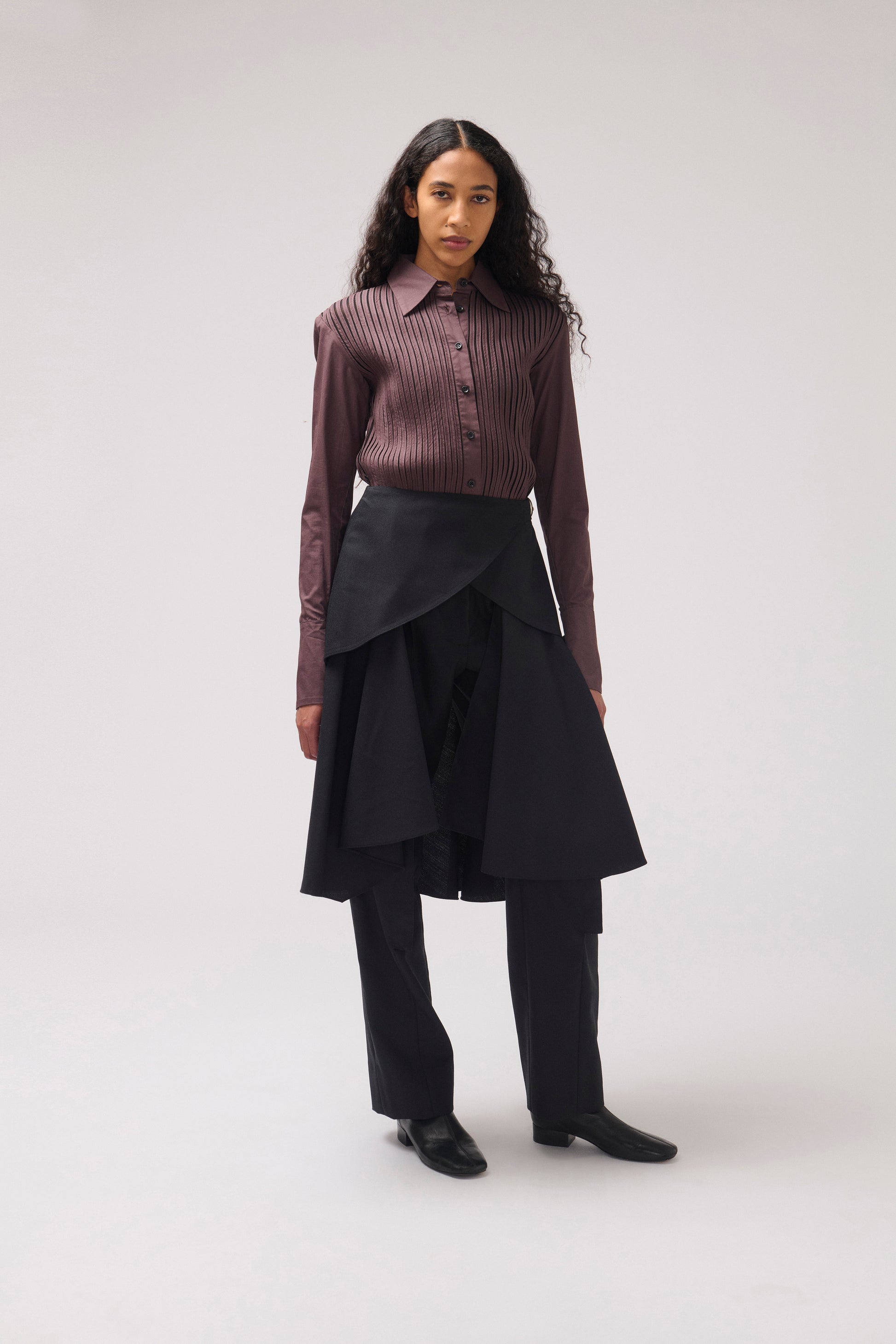 A model stands against a light gray studio background wearing a long-sleeve button-front shirt in a dark mauve-brown ribbed fabric, a black asymmetrical wrap skirt, black trousers, and black shoes.