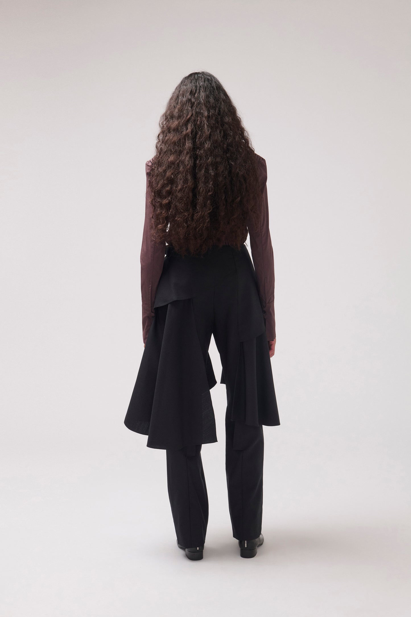 A model viewed from the back wearing a dark brown long-sleeve top and black high-waisted, layered asymmetrical trousers against a light gray studio background.