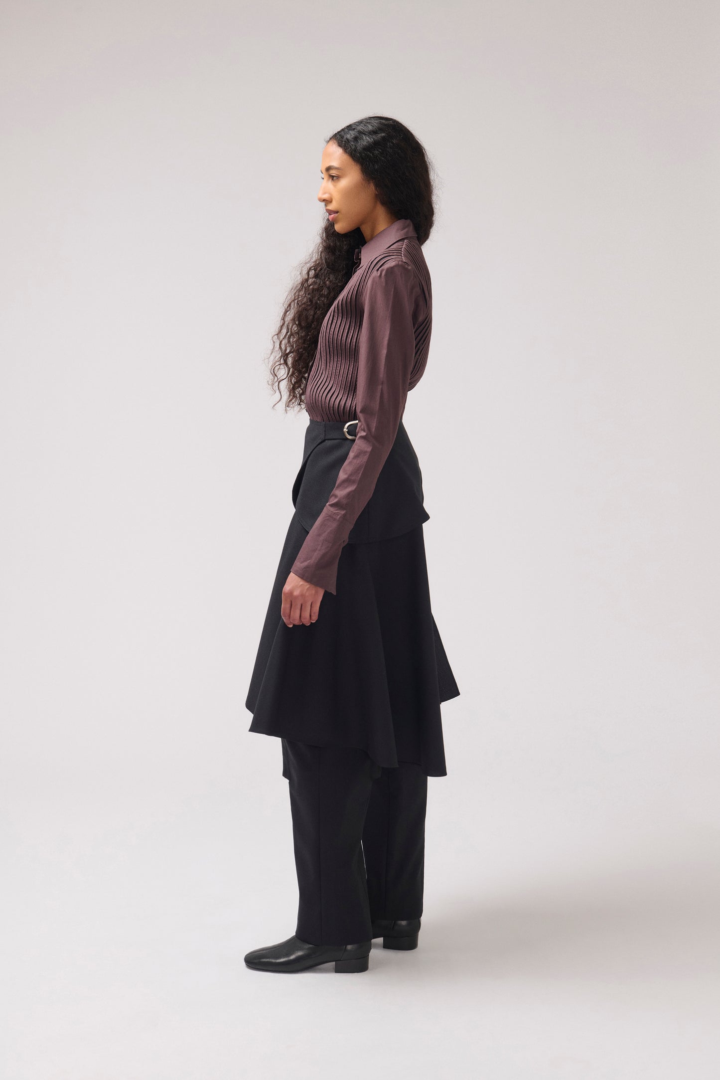 A woman stands in profile wearing a dark mauve ribbed long-sleeve top, a black layered skirt, black trousers, and black shoes against a light gray studio background.