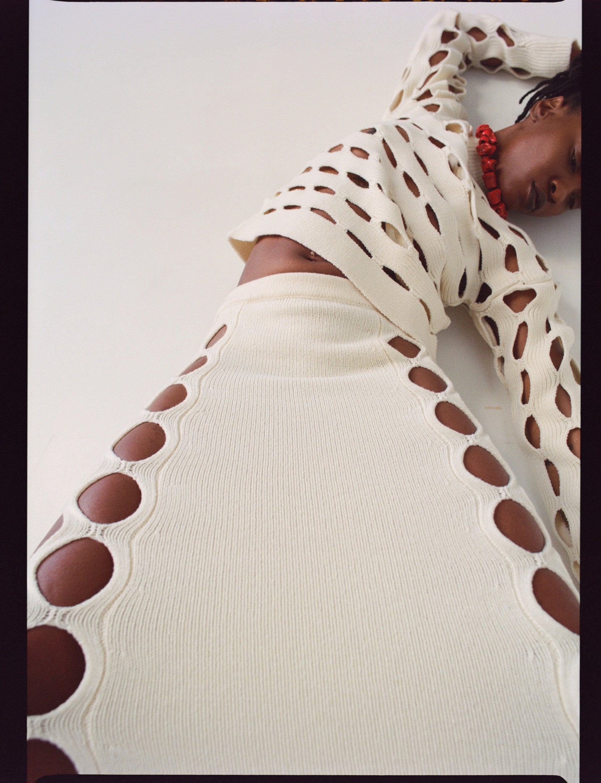 Model wearing a white knit matching set outfit with circular cutouts patterns on throughout the top and on the sides of the skirt styled with a chunky red beaa plain backgrounded necklace photographed against a white background 