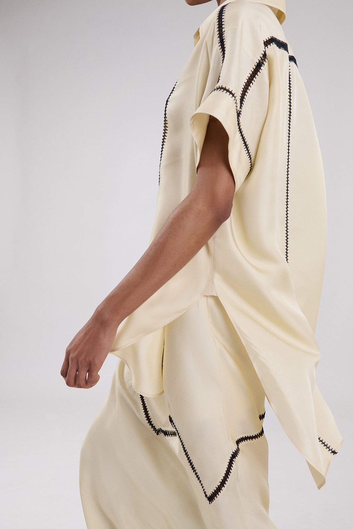 A close-up side view of a model wearing a cream-colored draped garment with black zigzag contrast stitching, set against a plain light gray studio background.