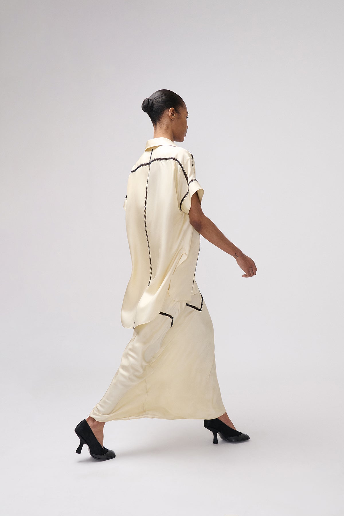 A model is shown from a rear three-quarter view walking while wearing a cream-colored, short-sleeved maxi dress with thin black piping details and black high heels, set against a plain light gray studio background.