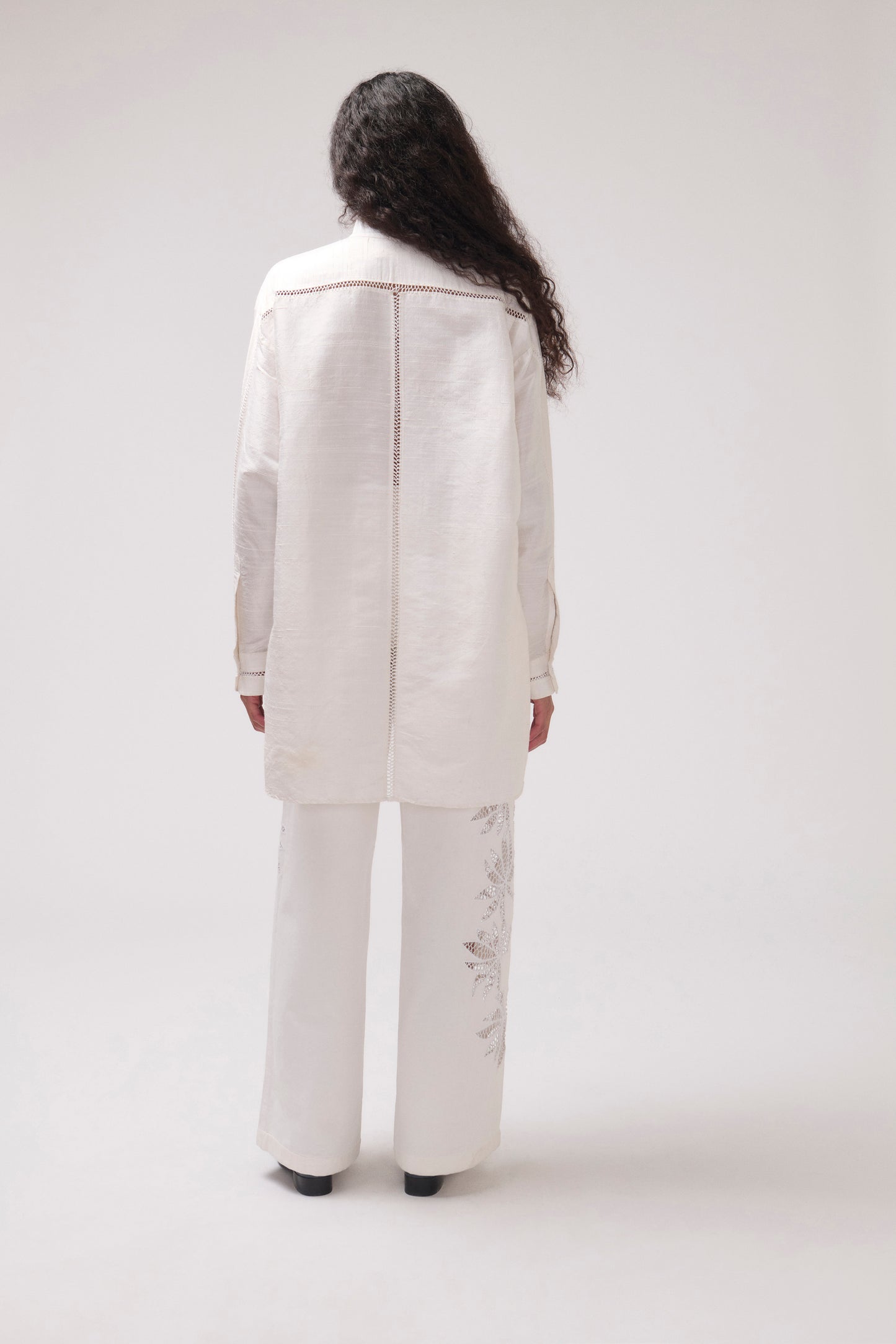 A model viewed from the back wearing a matching off-white two-piece set consisting of a long-sleeveshirt and wide-leg trousers with subtle embroidery on the lower legs, set against a plain light gray studio background.