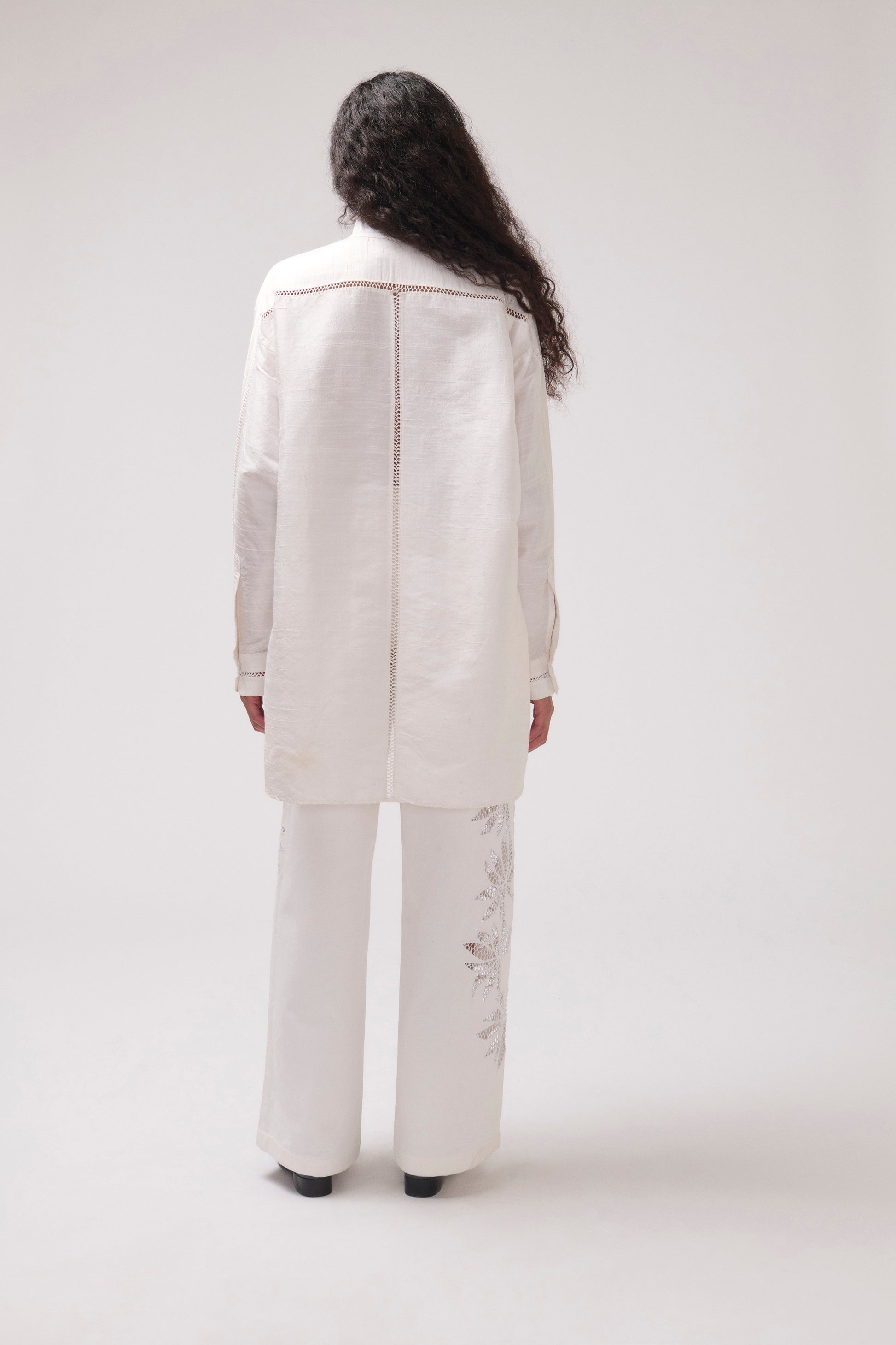 A model viewed from the back wearing a matching off-white two-piece set consisting of a long-sleeveshirt and wide-leg trousers with subtle embroidery on the lower legs, set against a plain light gray studio background.