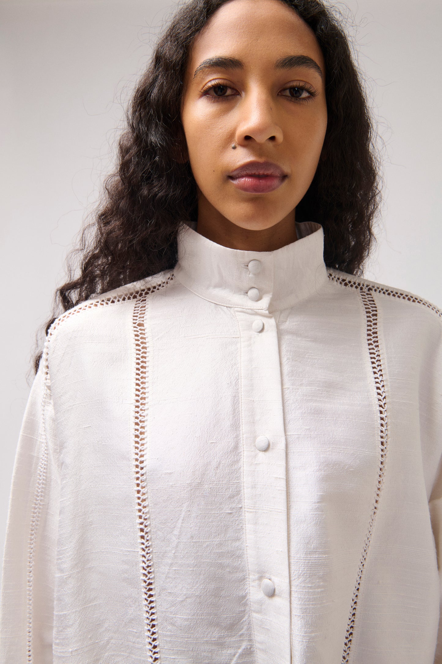 A model wearing a white high-neck button-up blouse with decorative vertical cutwork details on the shoulders and chest, photographed against a plain light gray background.