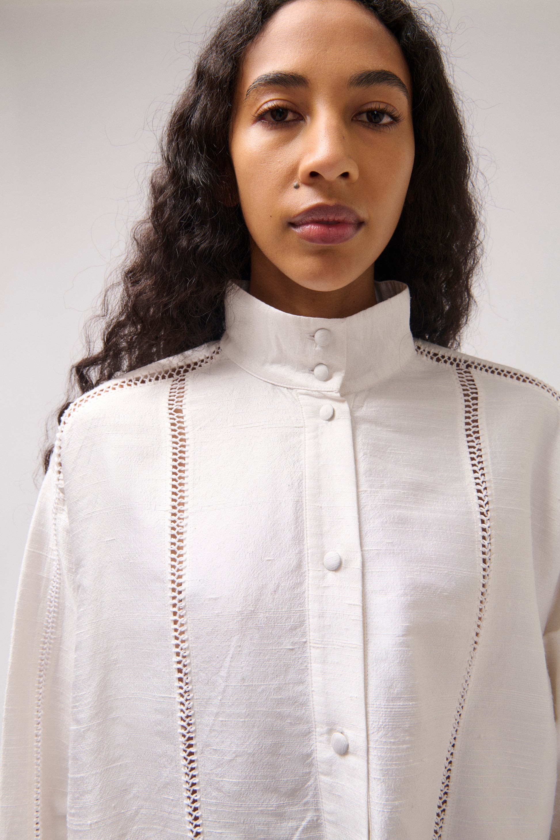 A model wearing a white high-neck button-up blouse with decorative vertical cutwork details on the shoulders and chest, photographed against a plain light gray background.