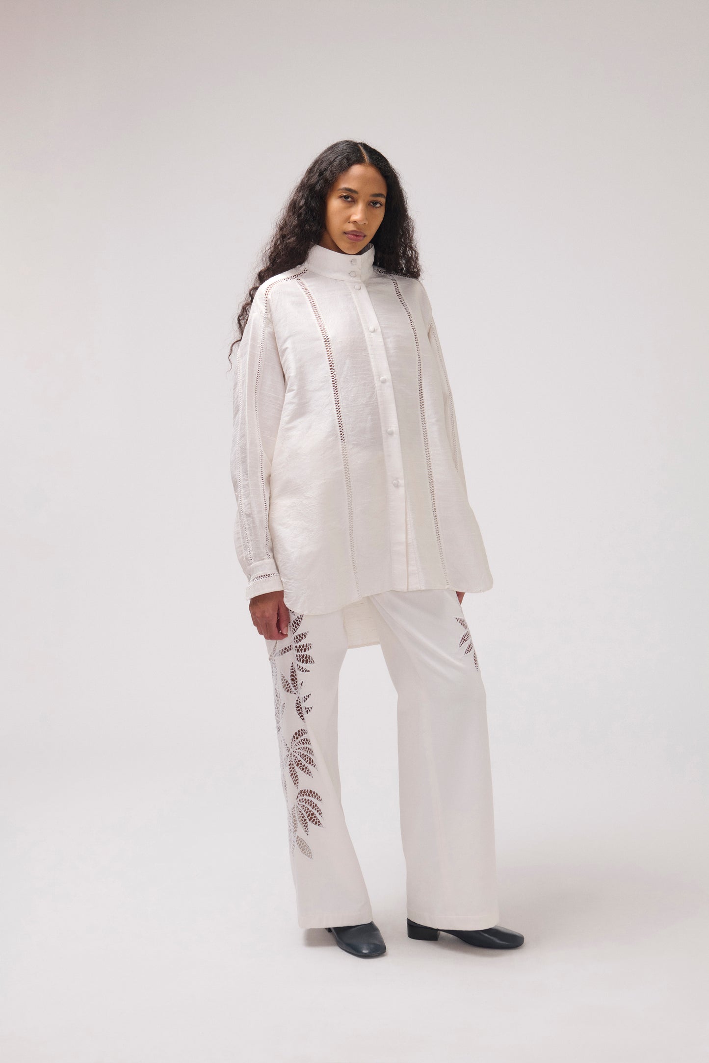 A model wearing a white two-piece outfit consisting of a long-sleeve button-up silk shirt and matching wide-leg trousers with cut-out leaf embroidery, photographed against a light gray studio background.