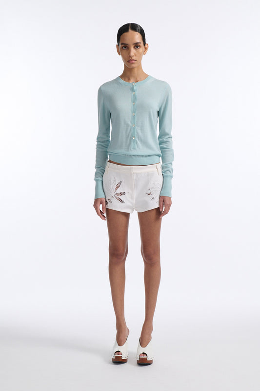 Female model with slicked back black hair in a bun wears blue fine gauge long sleeve button down cardigan and white short shorts with palm leaf cutouts at front and back.