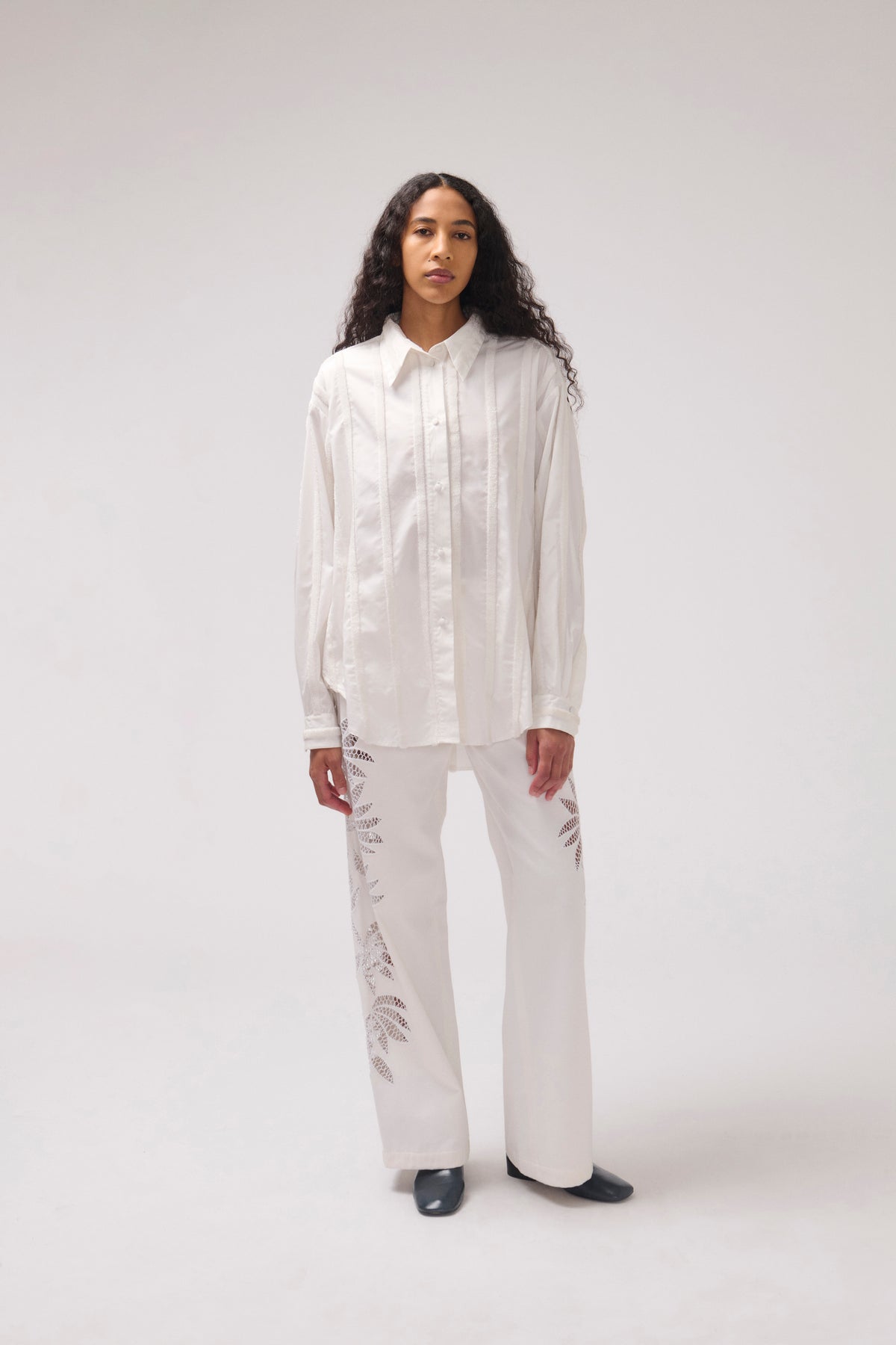 A model stands against a light gray studio background wearing a white long-sleeve button-up shirt and matching white wide-leg pants with subtle tonal leaf embroidery near the lower legs.