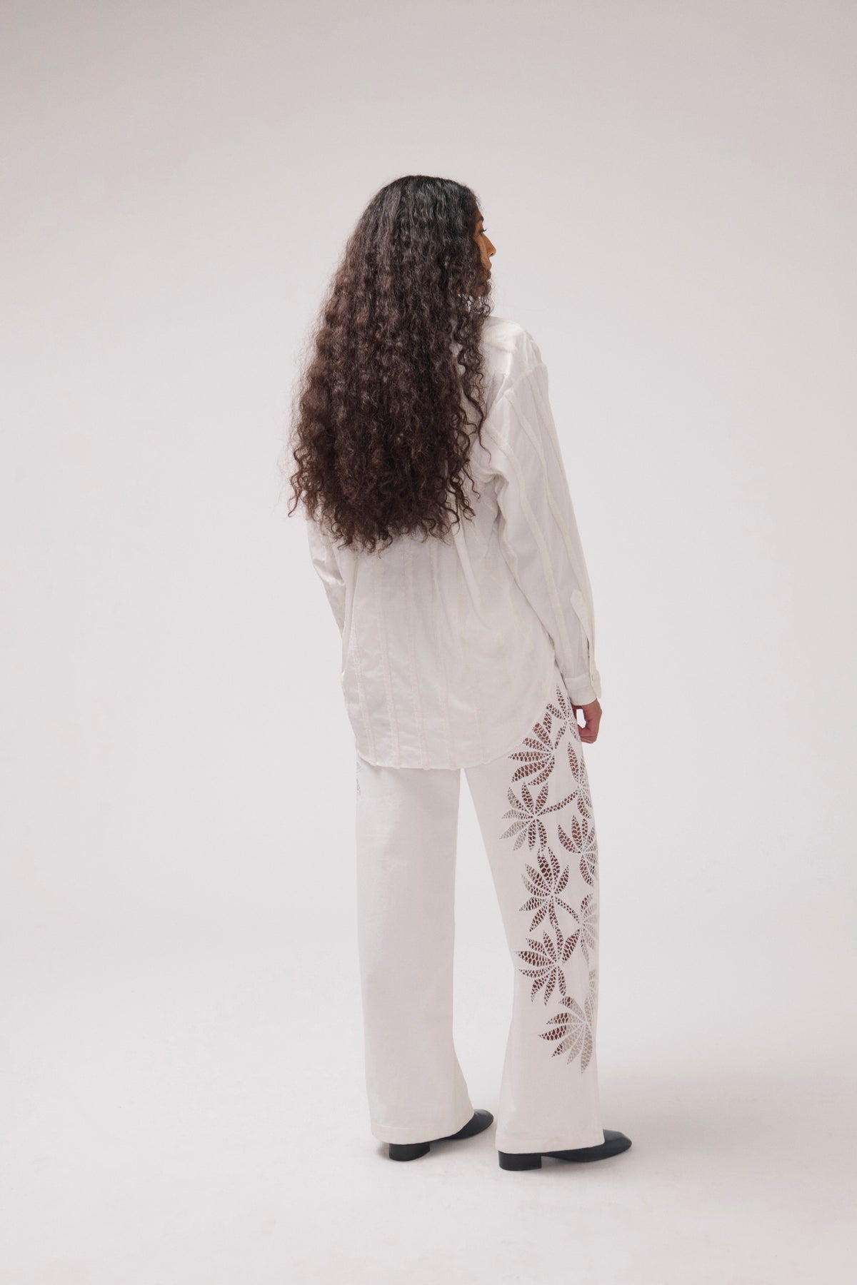 A woman with long, curly dark hair is shown from behind wearing a white long-sleeve shirt and matching wide-leg pants with brown leaf embroidery down the side of the pants, set against a plain light gray studio background.