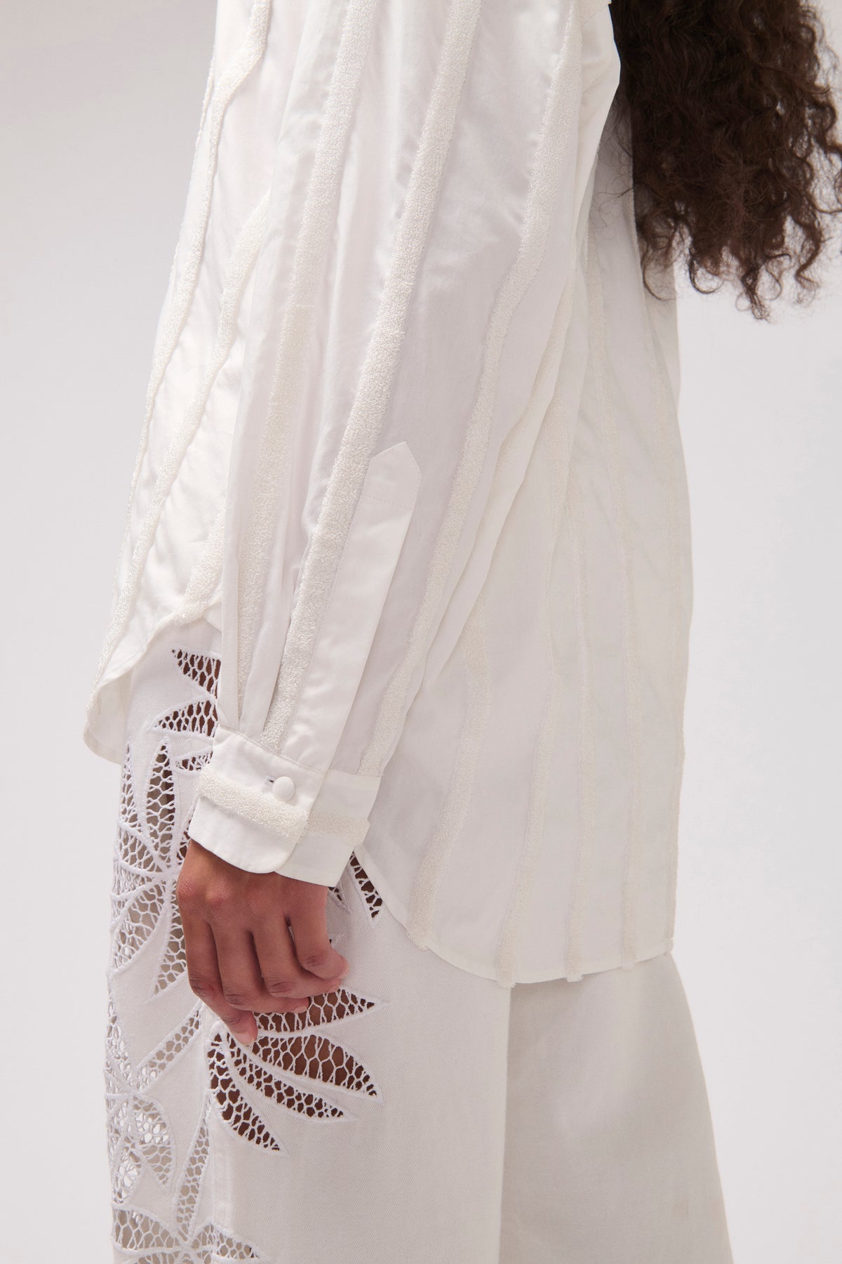 A close-up of a person wearing a white long-sleeve blouse with sheer floral eyelet lace panels along the lower sleeves and a matching white skirt or trousers, photographed against a plain light background.