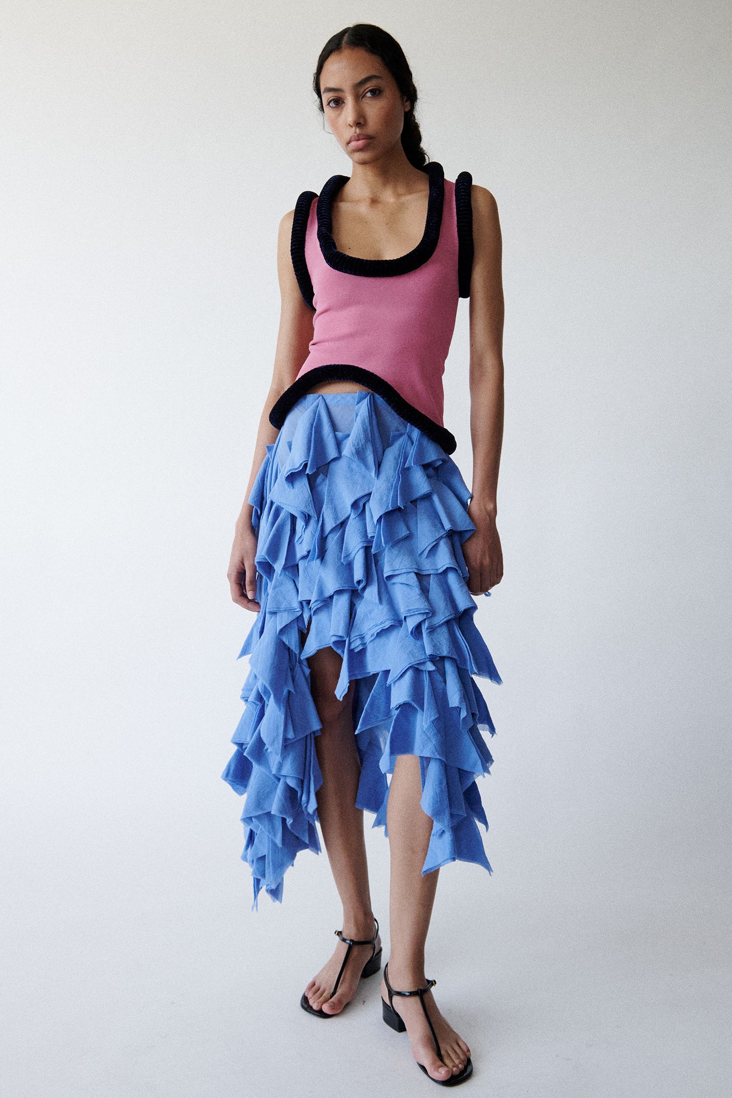 Model wears pink tank top with sculptural piping and blue asymmetric ruffle skirt with thin black thong sandals on a white background. 