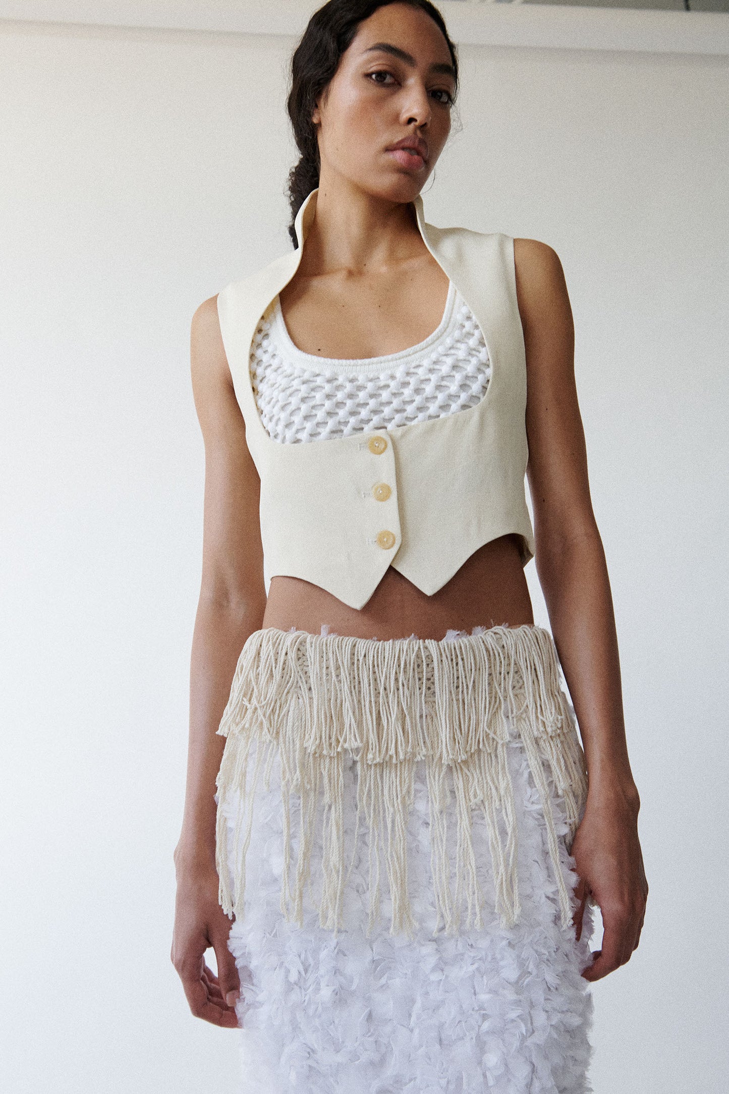 Model wears cream colored open neckline vest with white crystal embellished bralette and white fluffy textured skirt. 