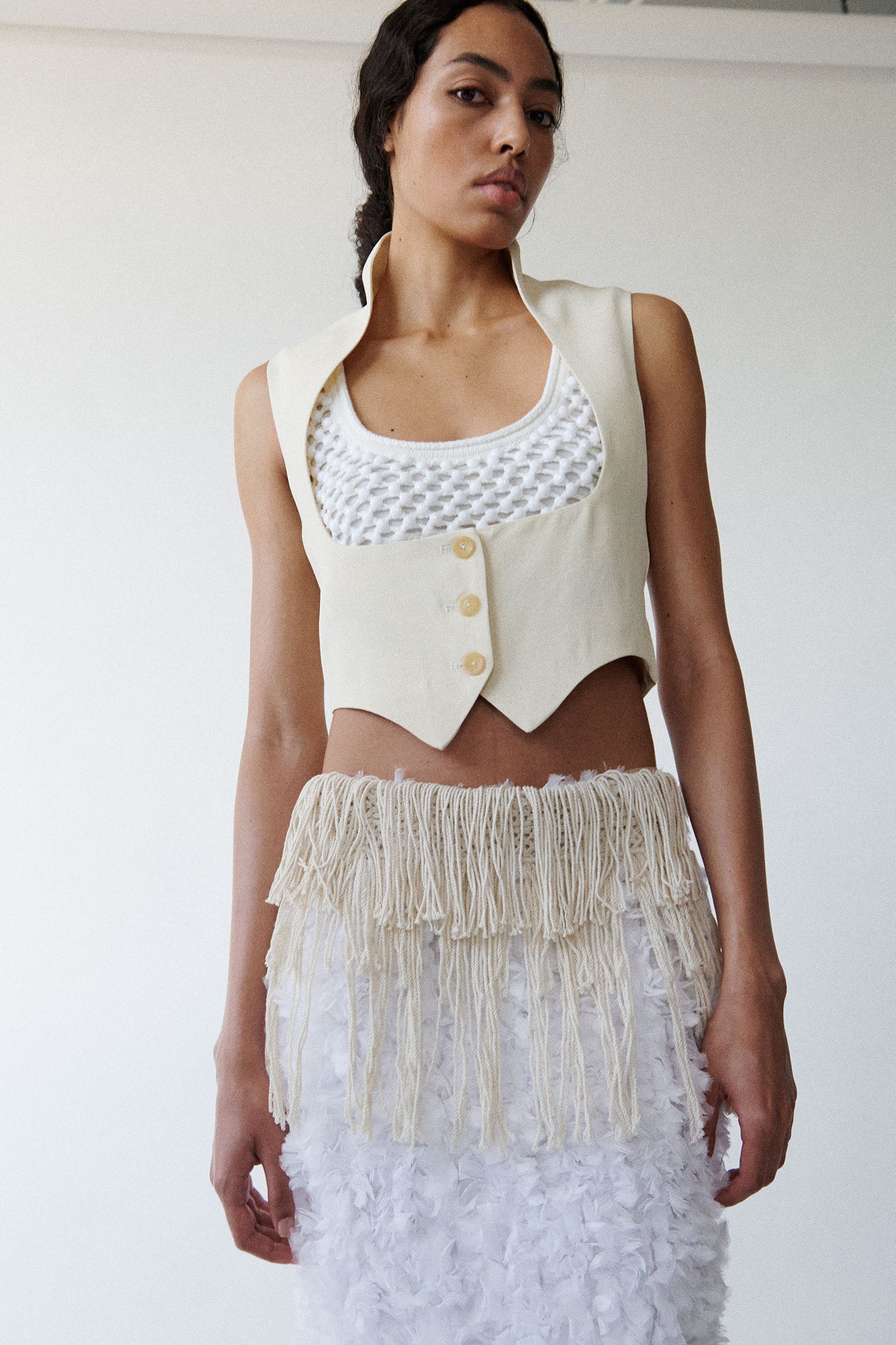 Model wears cream colored open neckline vest with white crystal embellished bralette and white fluffy textured skirt. 