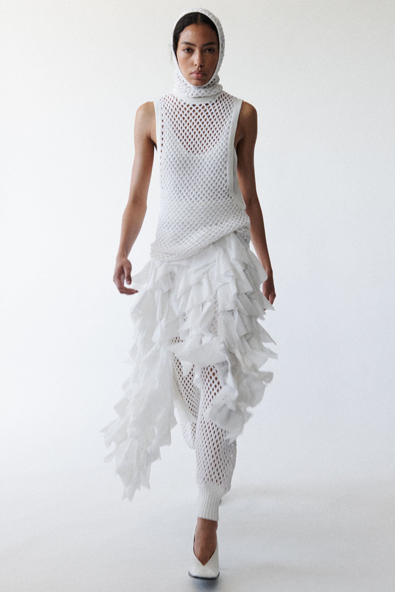 Model wears white balaclava hood, long sleeve crewneck sweater, and elastic waisted trousers. All pieces are unlined fine mesh embellished with small crystals. Model also wears white asymmetric ruffle skirt and white pumps. 
