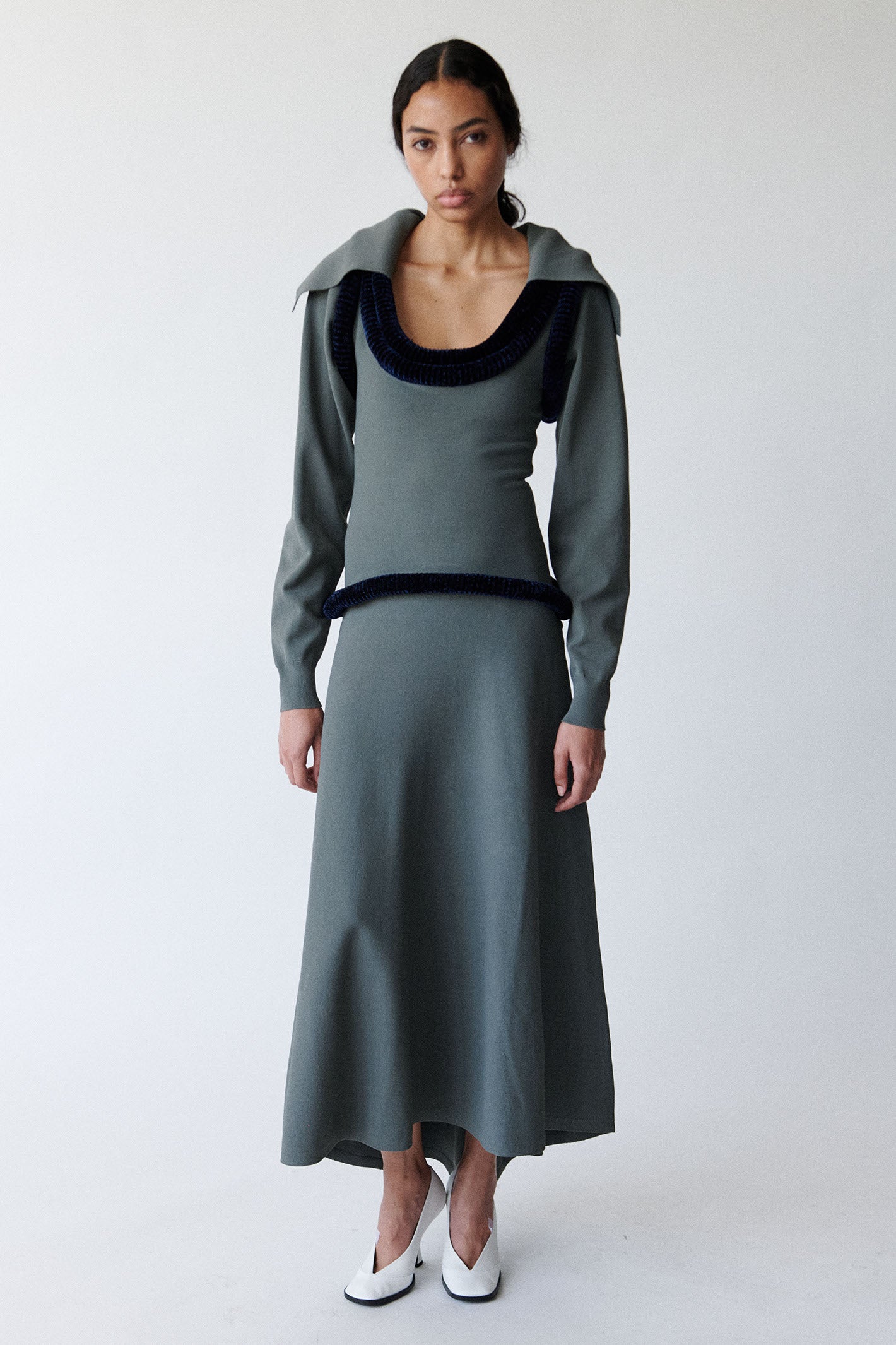 Model is wearing a gray dress with black pipping and a gray collar shirt layered underneath paired with white pumps photographed on a white background