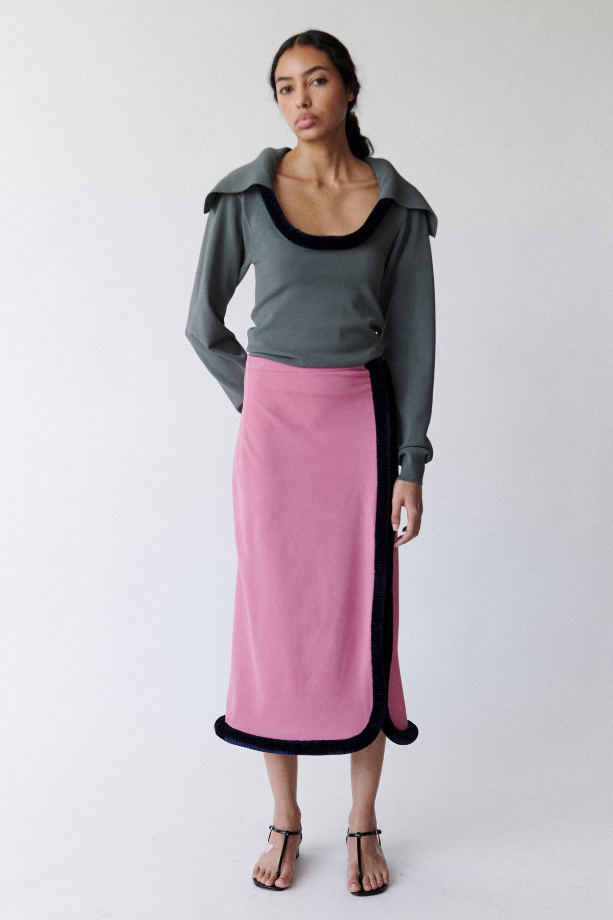 Model wearing cool-toned grey long sleeve sweater with sculptural piping along the neckline and a draped sailor's collar with a bubble-gum pink long skirt with sculptural piping along the front and hem. 