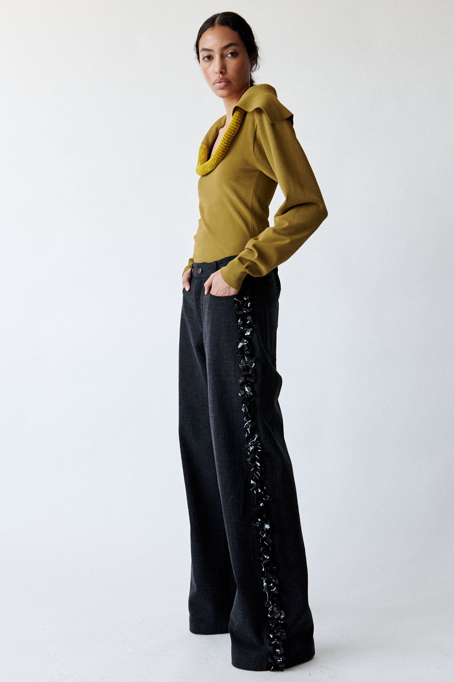 Model wears cumin colored knit sweater with sculptural piping and black denim trousers with black paillette embellishments on both legs. 